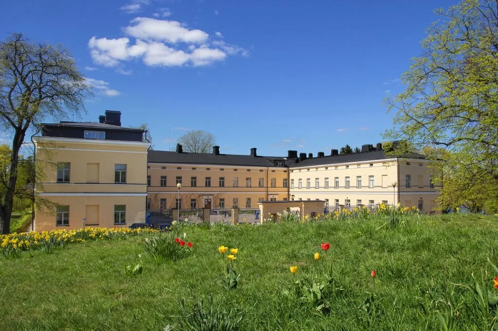 A park with green grass and blooming flowers in front of multi-story residential buildings under a blue sky with some clouds.