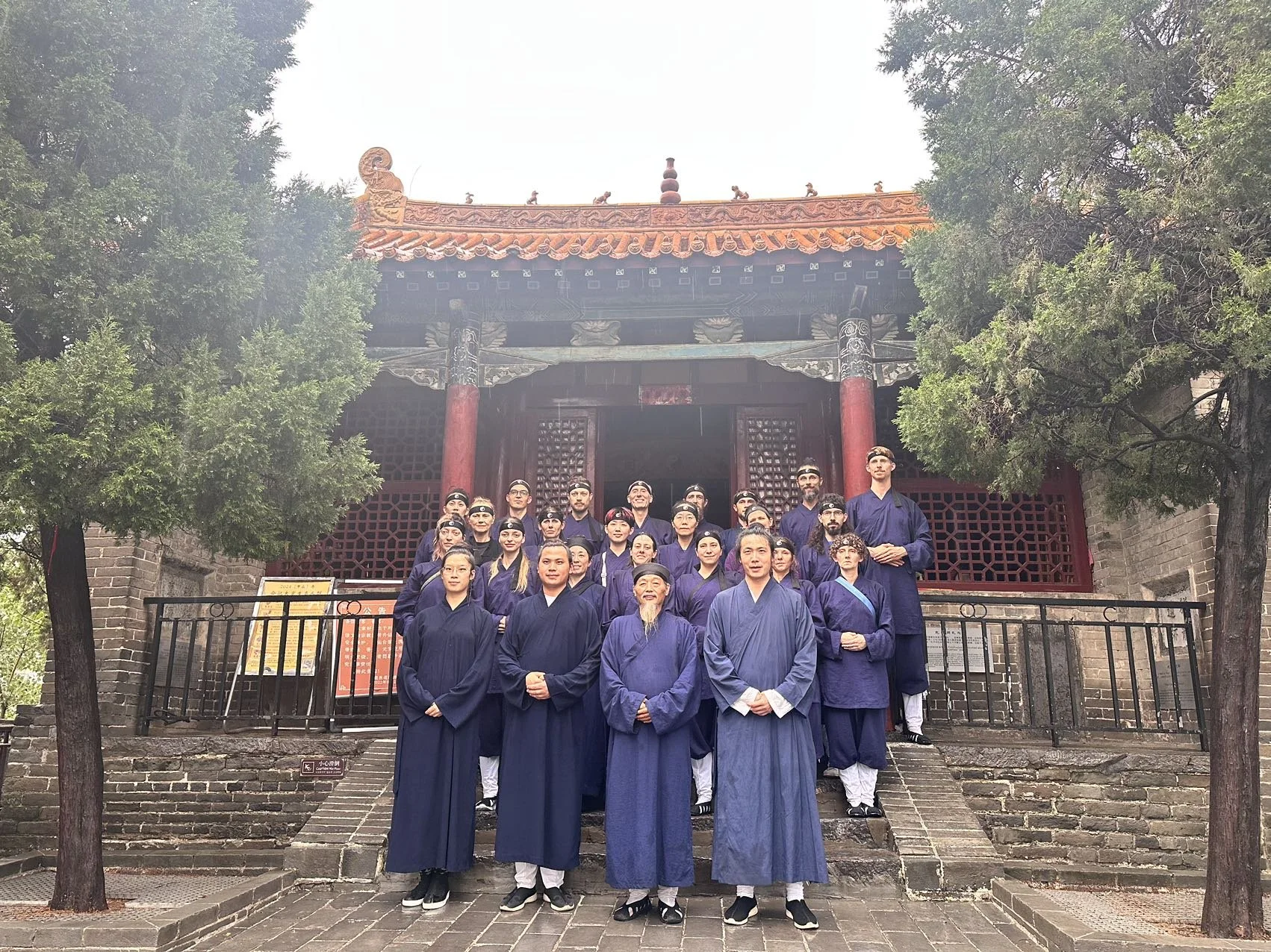 Group of people wearing traditional blue Chinese clothing standing on stone steps in front of a historic Chinese building with red columns and ornate roof, flanked by trees.