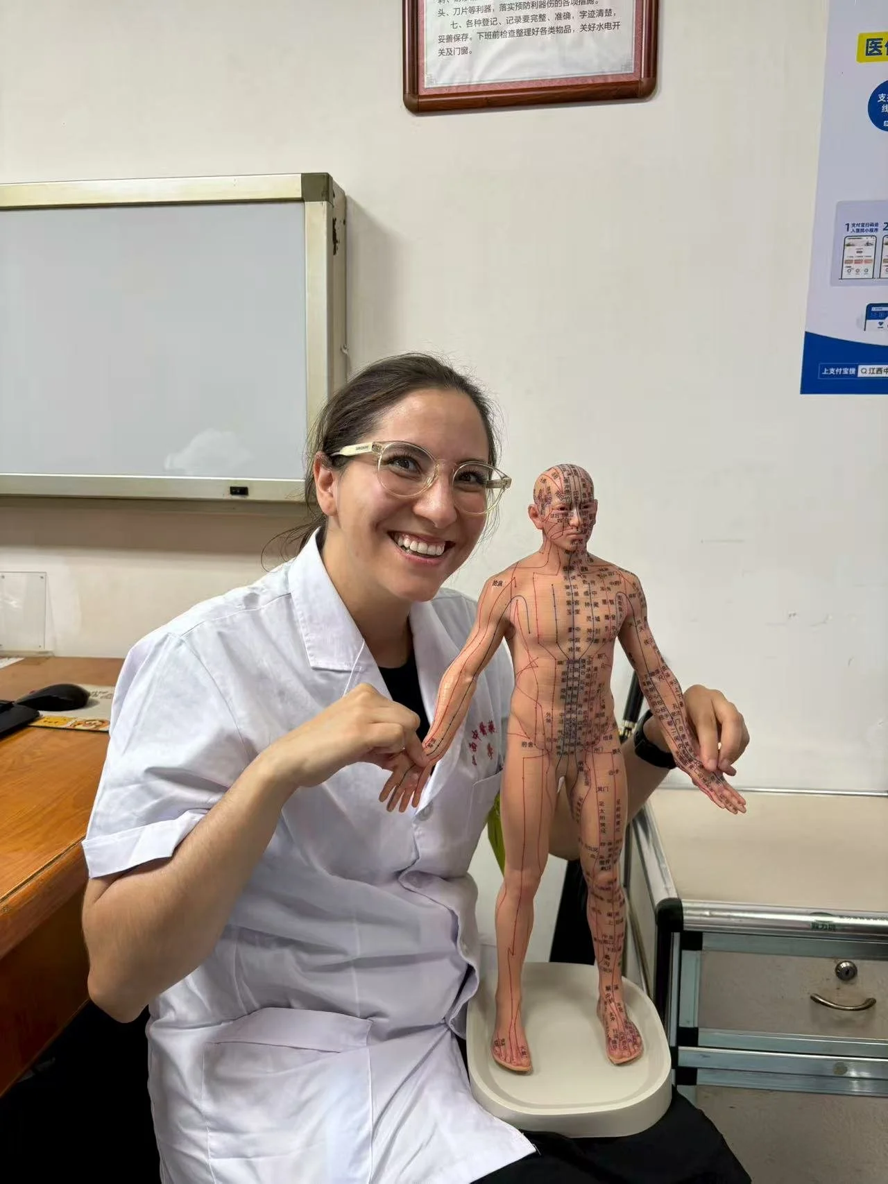 A woman in a white coat holding a traditional Chinese medicine acupuncture model, smiling in a medical or clinic setting.