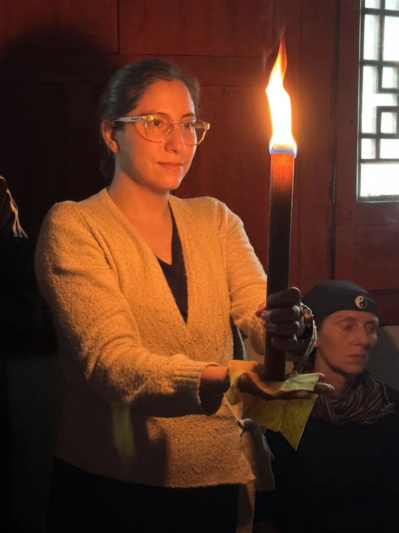 Woman holding a lit candle inside a dimly lit room with wooden walls and a window.