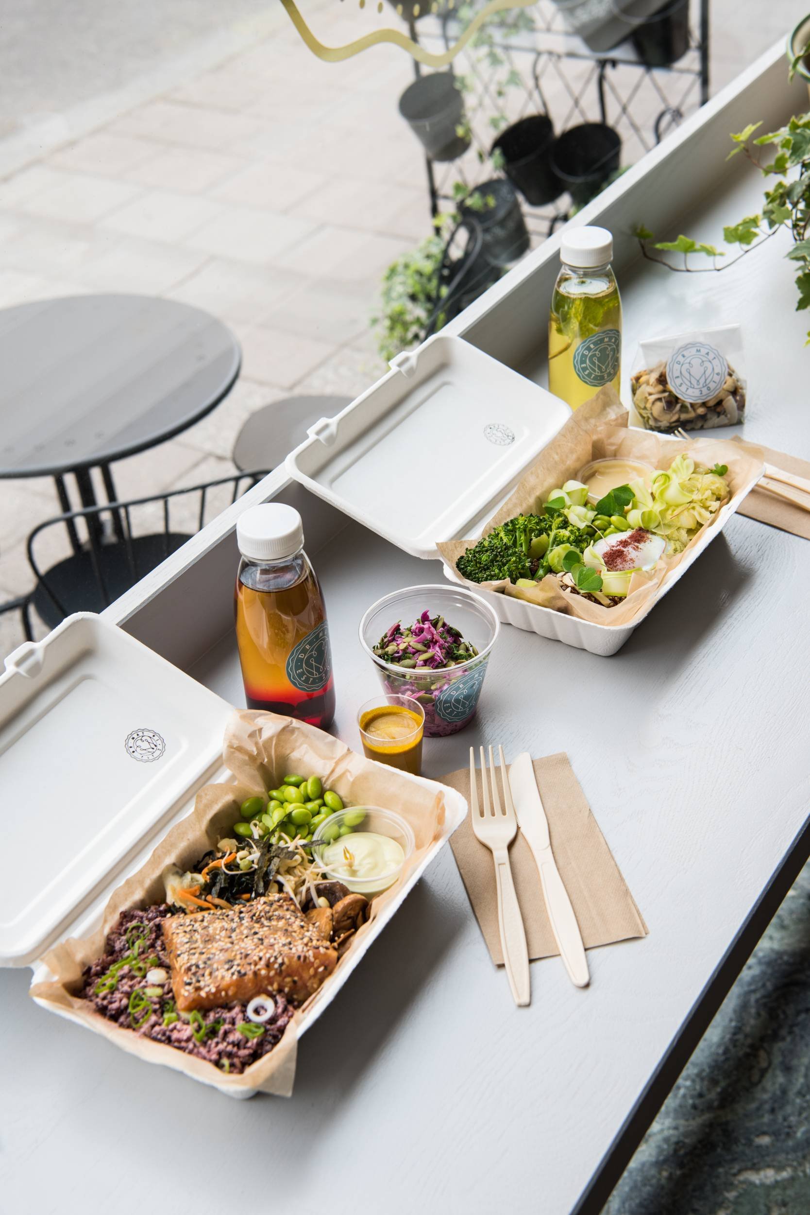 Takeout meal with salads, tofu, grapes, drinks, and condiments on a gray table outdoors.