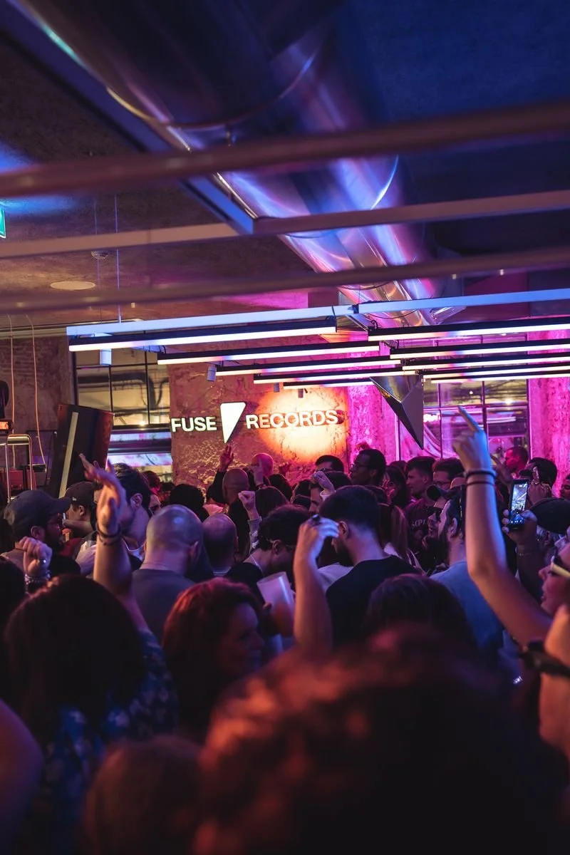 Crowd of people dancing and enjoying music at Fuse Records event with neon lights and a illuminated sign.