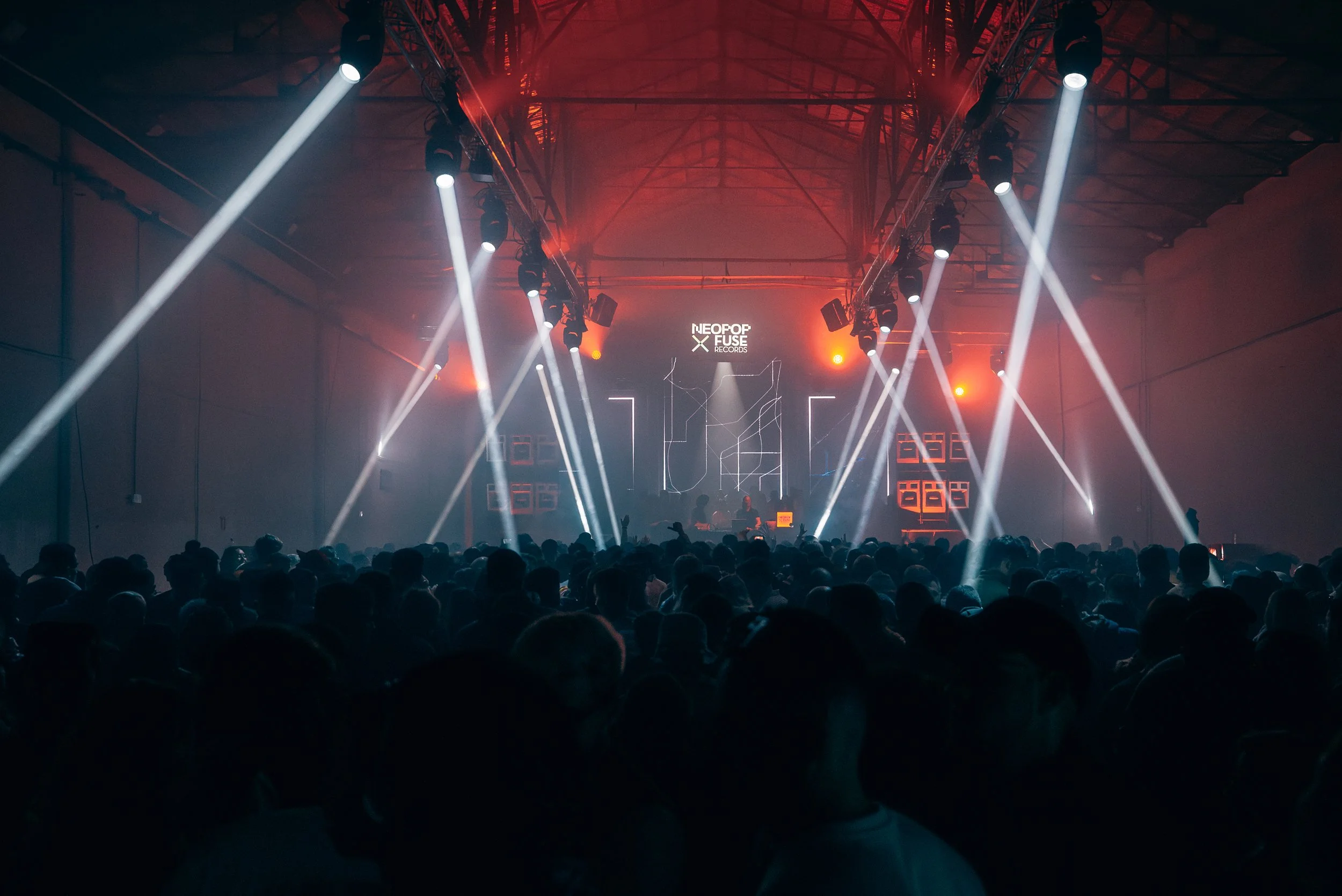 Crowd at a music event with neon lights and speakers, featuring a sign for Neopop Fuse Records.