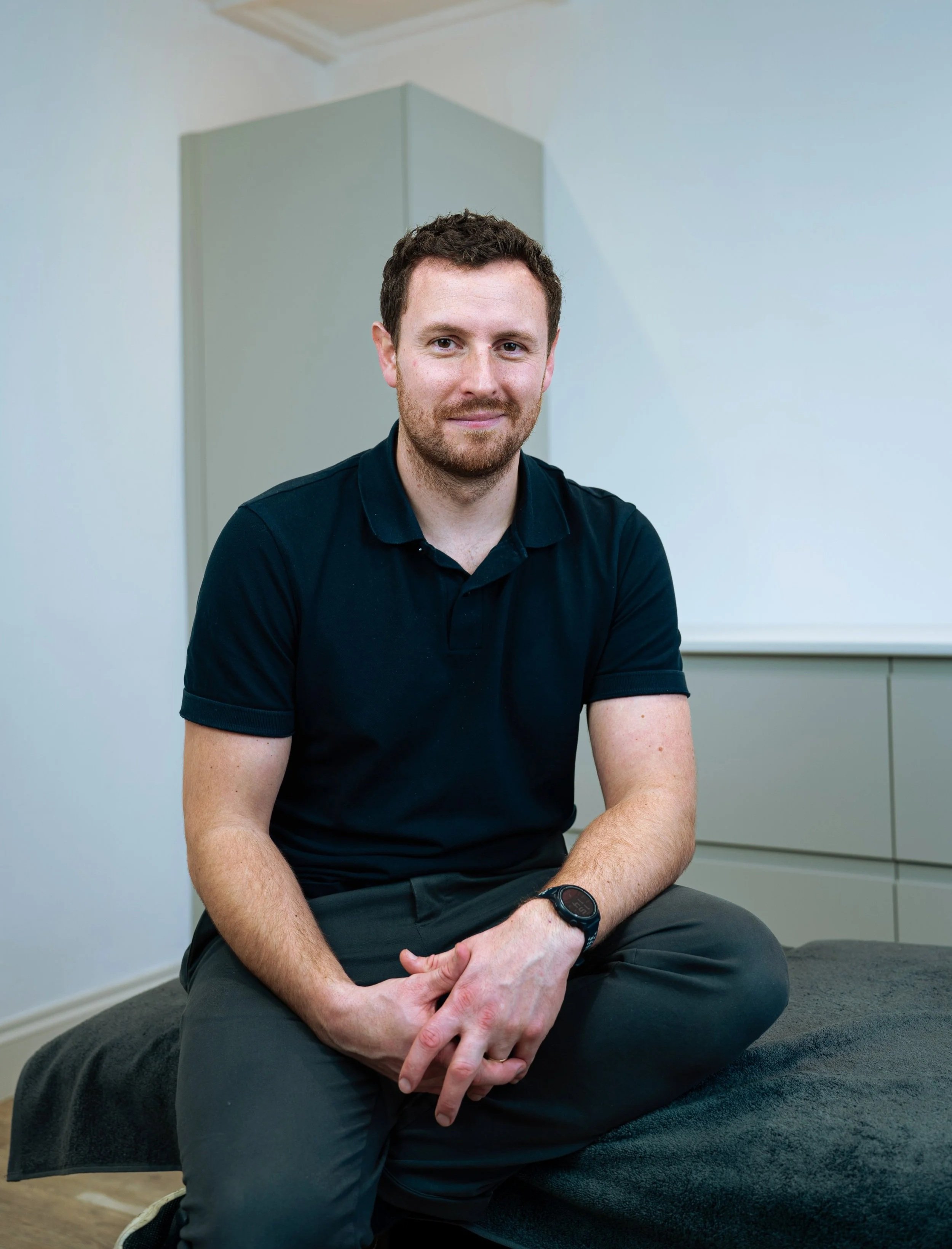 Rob Bale, Physiotherapist and owner of Revive Physio Cornwall seated on a treatment table in clinic.