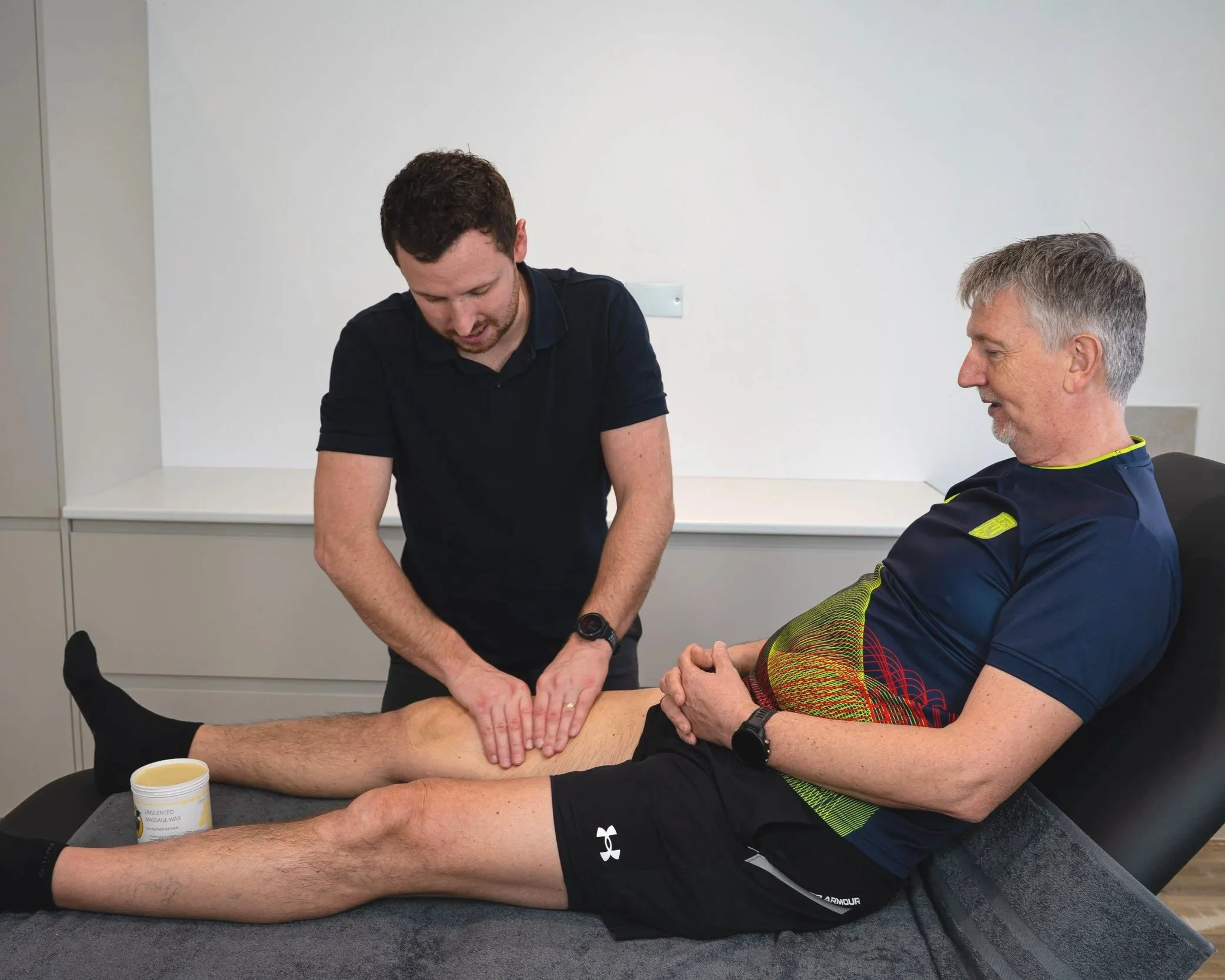 Rob Bale Physio performs a massage or therapeutic manipulation on a man's leg while the man sits on a treatment table in a clinical setting.