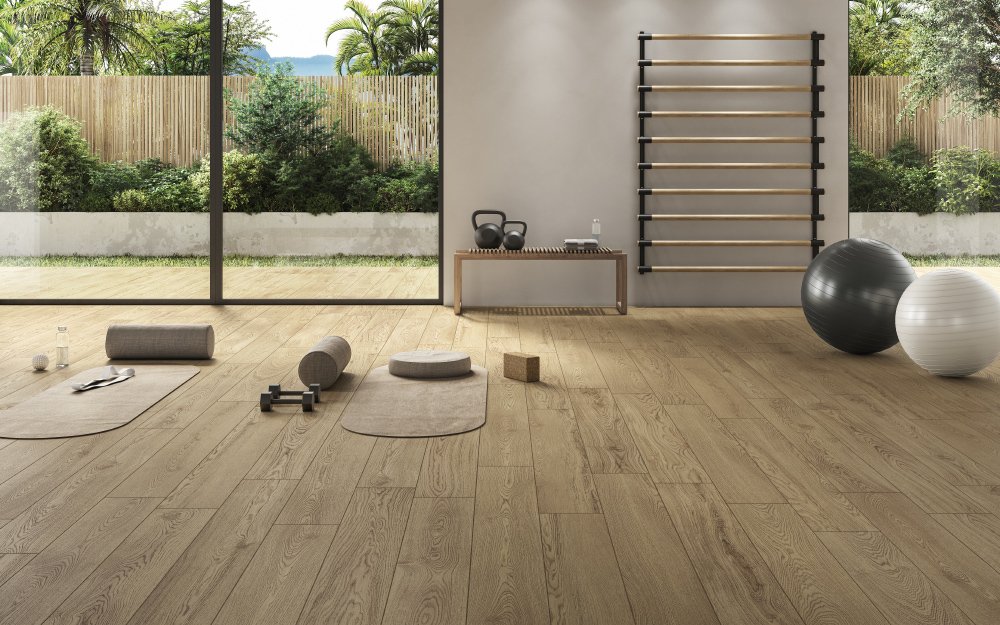 Heller Fitnessraum mit Holzboden, Yogamatten, Gymnastikbällen, Hanteln und Fitnesszubehör vor einer großen Fensterfront mit Blick auf einen grünen Garten.
