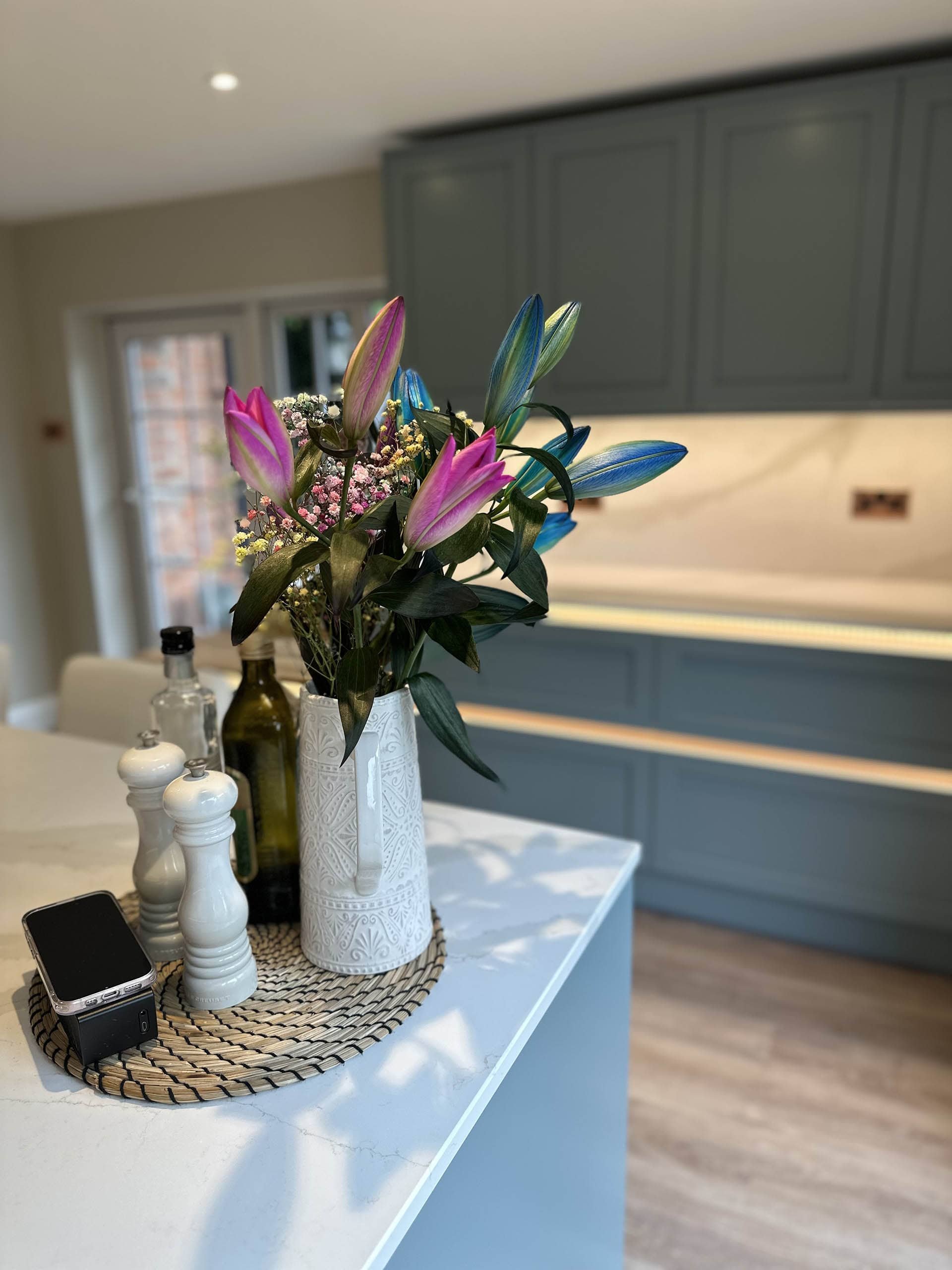 Modern kitchen with a white countertop featuring a decorative floral arrangement in a patterned vase, surrounded by a black pepper mill, salt grinder, a bottle of olive oil, and a smartphone. Gray cabinetry is visible in the background.
