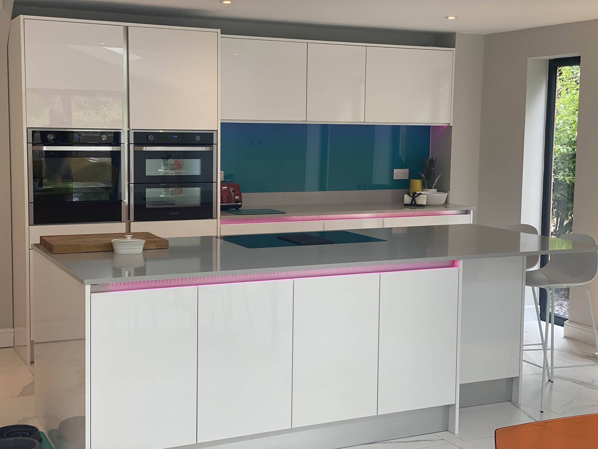 Modern kitchen interior with white cabinets, built-in ovens, and island with a cooktop. Pink LED lights under the countertop and blue backsplash add color. A window provides natural light.