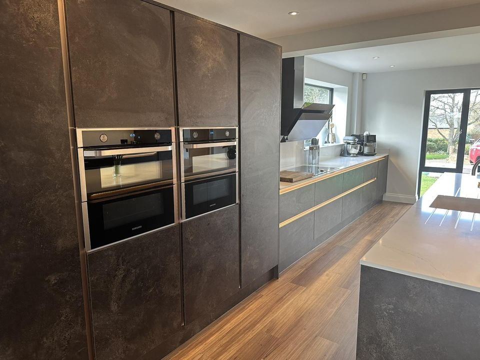 Modern kitchen with dark cabinetry, built-in ovens, stainless steel appliances, a countertop with a sink, and large windows.