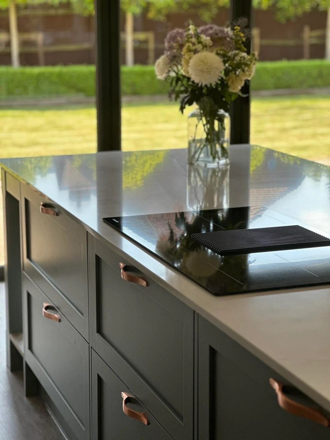 Kitchen island with black cabinets, copper handles, an induction cooktop, and a vase of flowers on a countertop overlooking a garden.