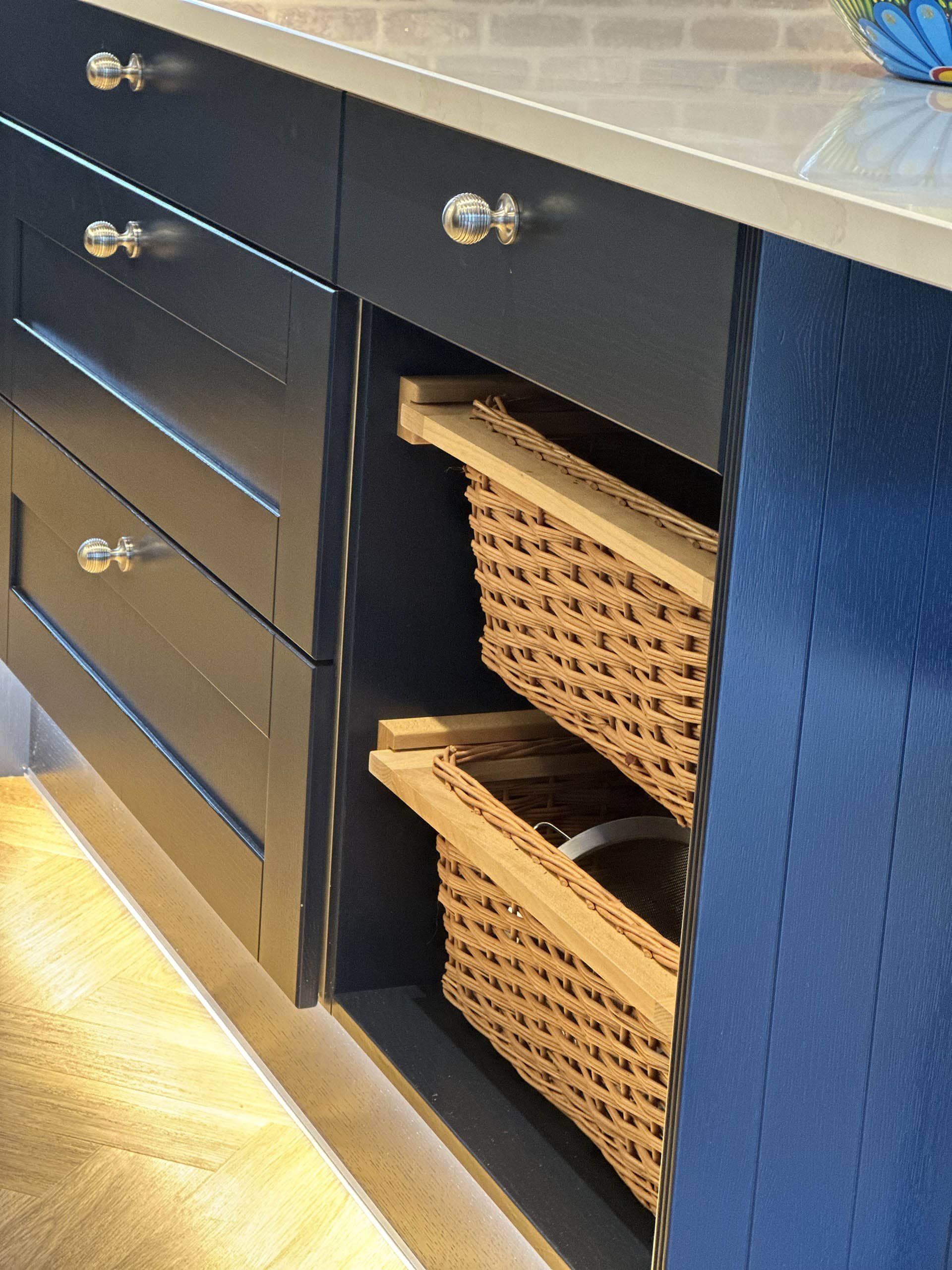Dark blue kitchen cabinet with silver knobs and wicker baskets in pull-out drawers under a light countertop.