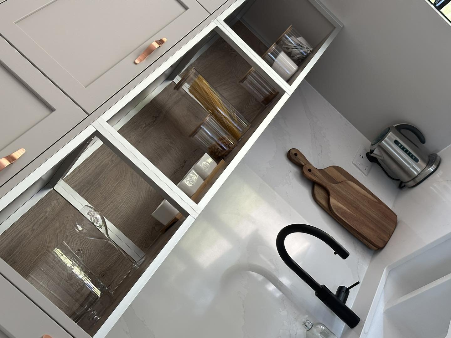 Kitchen corner with light gray cabinets, shelves with glass containers, electric kettle, black faucet, cutting boards, and a white countertop.