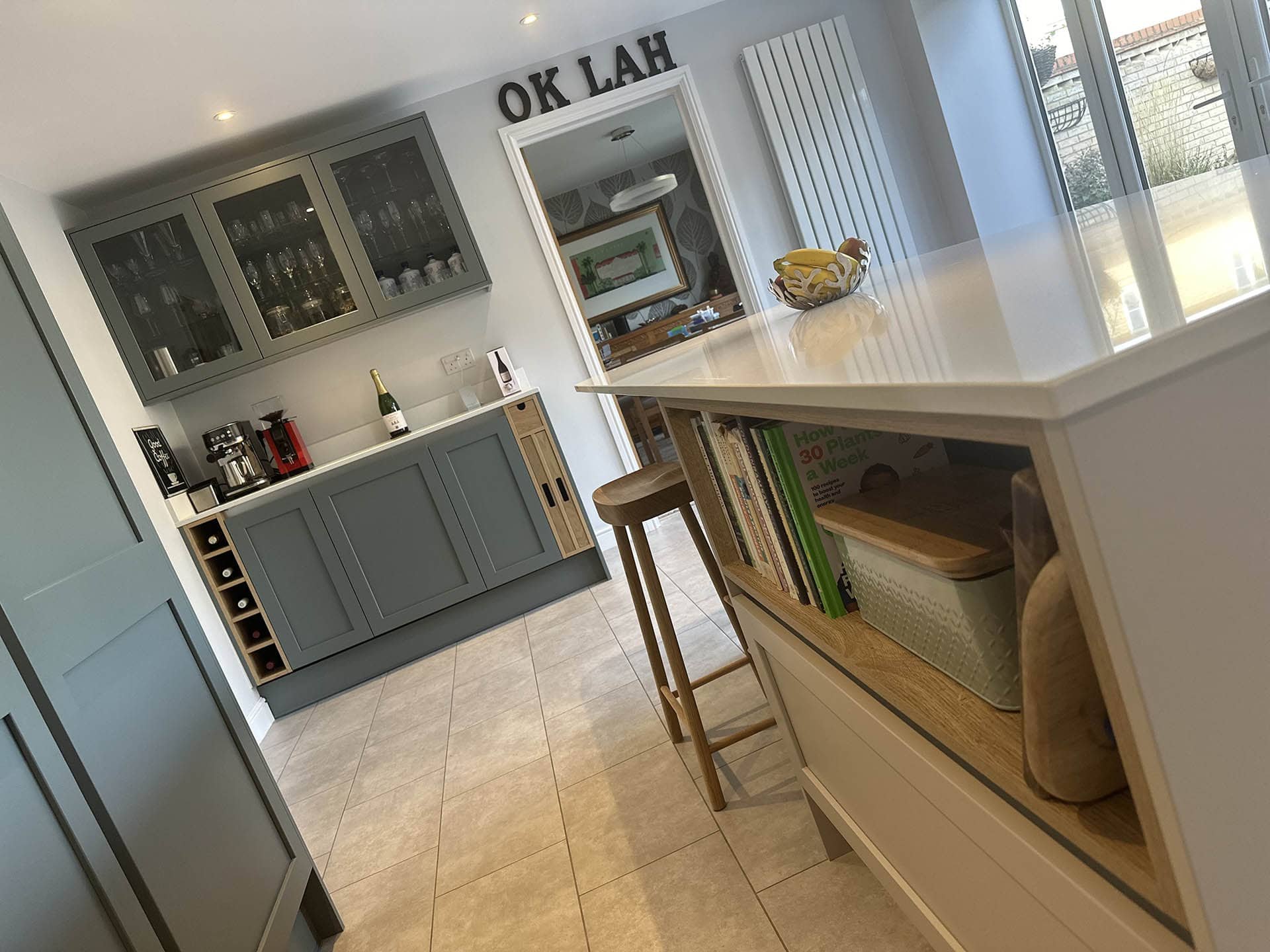 Shaker kitchen interior with green cabinets, a coffee machine, wine rack, and cookbook shelf. A countertop holds a champagne bottle and fruit bowl. Natural light enters through a glass door. A stool is at the counter.