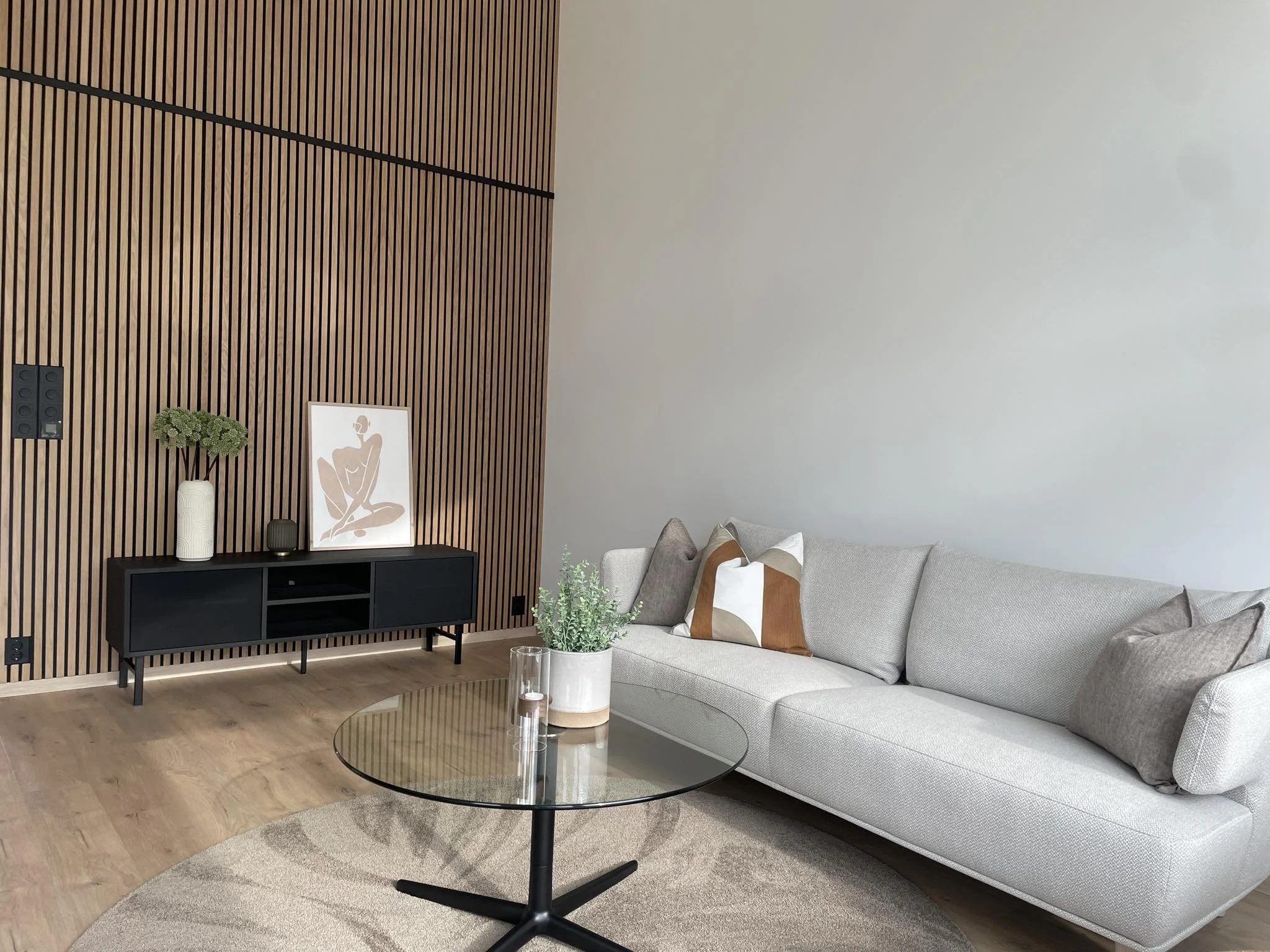 Moderne stue med grå sofa, rundt glassbord, og en vegg med trepanel. Dekorative puter på sofaen og bøker og plante på TV-bordet. Lyst tregulv og minimalistisk interiørdesign.