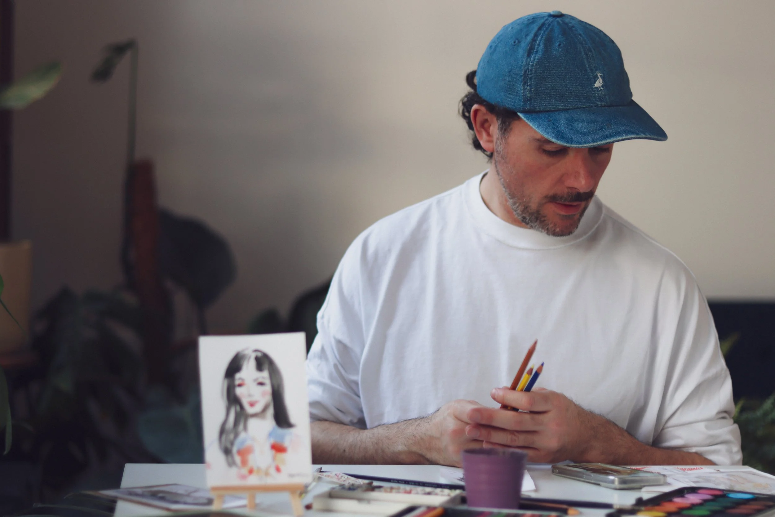 Eduard Illustrating , sitting at a table with art supplies, looking down while holding coloured pencils.