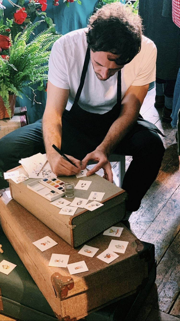 A man with dark hair and a beard, wearing a white t-shirt and black apron, is sitting on a wooden table while painting small cards with watercolor paints. The table has scattered cards with various colorful illustrations and a box of watercolor paints. There are vibrant red flowers and green fern plants in the background, creating a lively atmosphere.