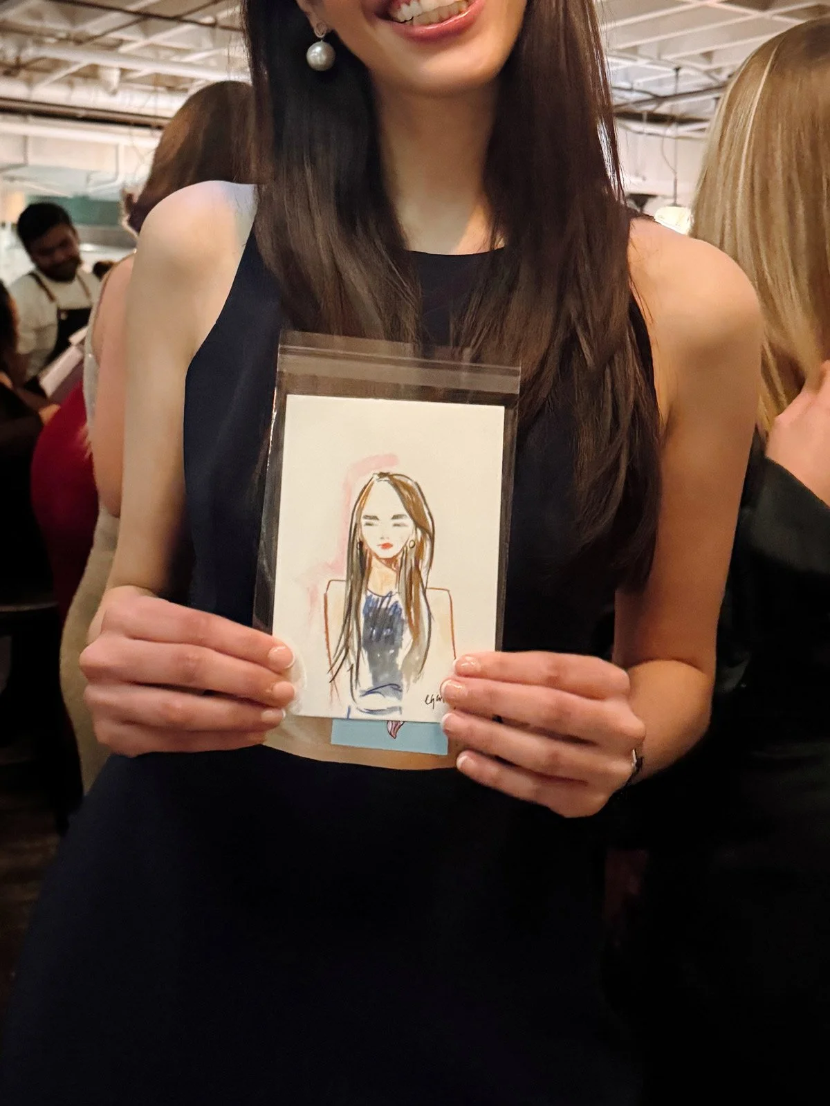 Woman in black dress holding a small drawing of herself, smiling in a crowded indoor setting.