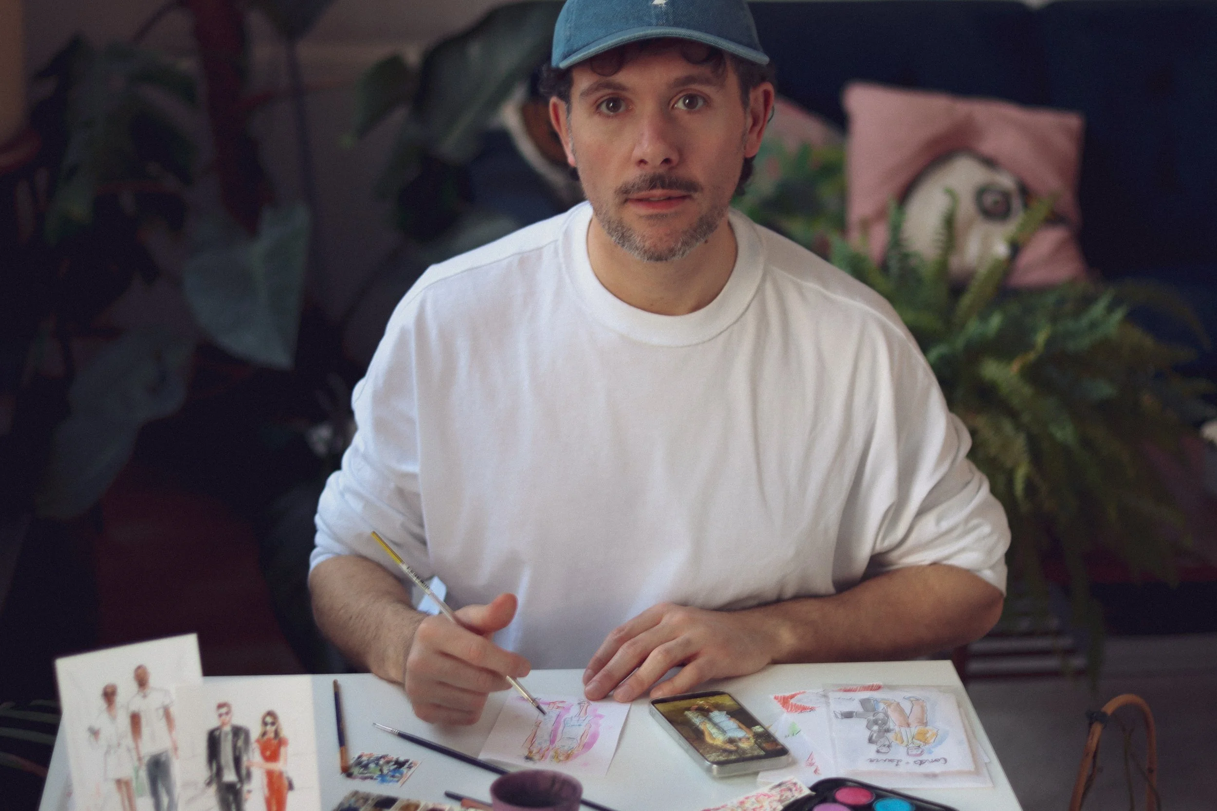 Eduard illustrates wearing a white shirt and a blue cap sitting at a table with watercolour paintings, brushes.