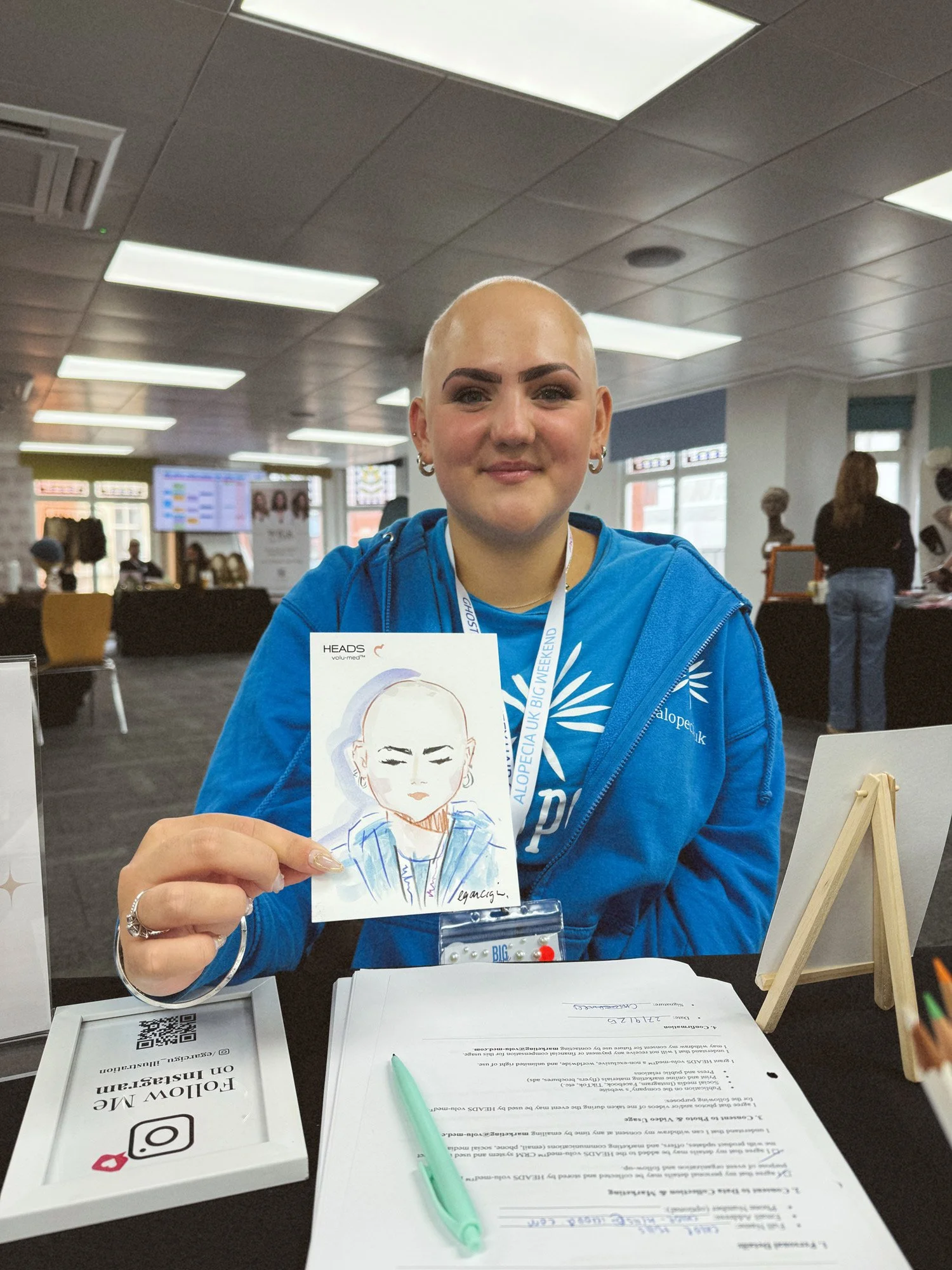 A woman with a shaved head and a blue jacket sits at a table in a busy indoor event, holding a drawing of herself by Eduard Illustrates