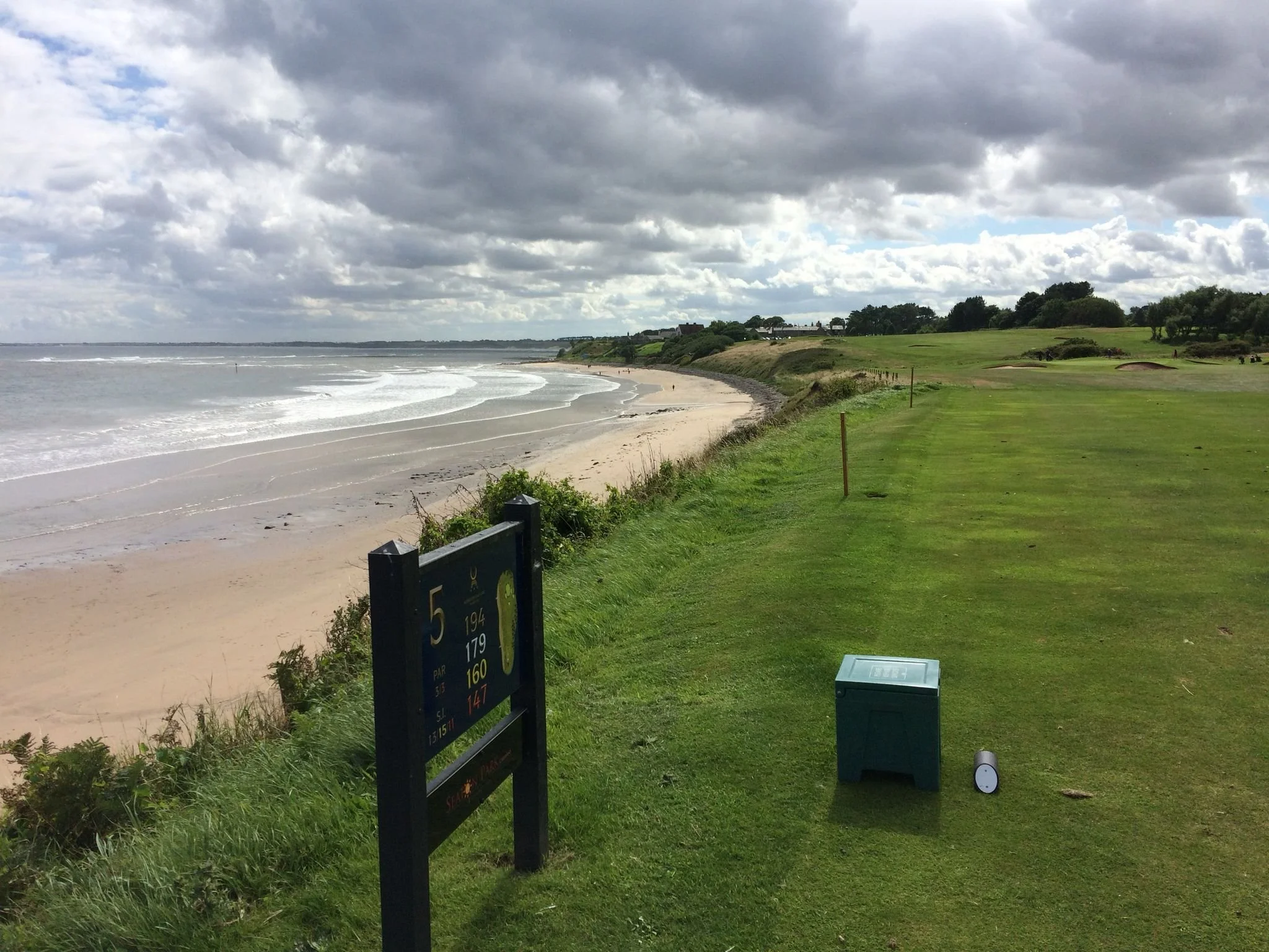 Golf course with a seaside view, including a green fairway, sandy beach, ocean waves, and a cloudy sky.
