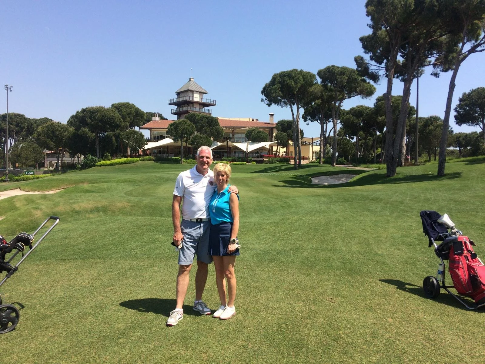 Man and woman standing on a golf course with trees and a clubhouse in the background, golf bags visible on the grass.