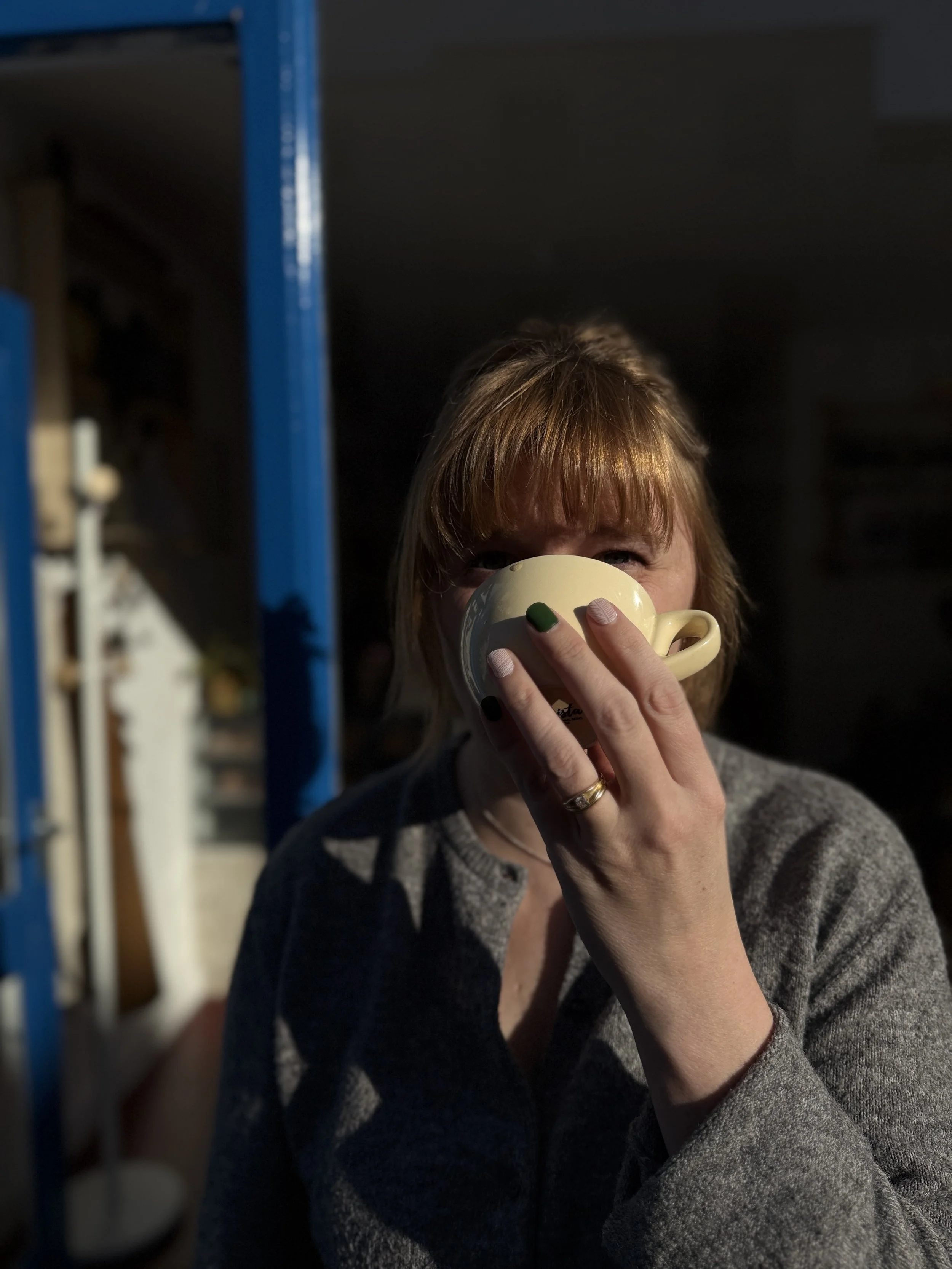 A woman with light brown hair and bangs drinking from a yellow mug with green and pink nails, wearing a gray sweater, standing outdoors in sunlight.