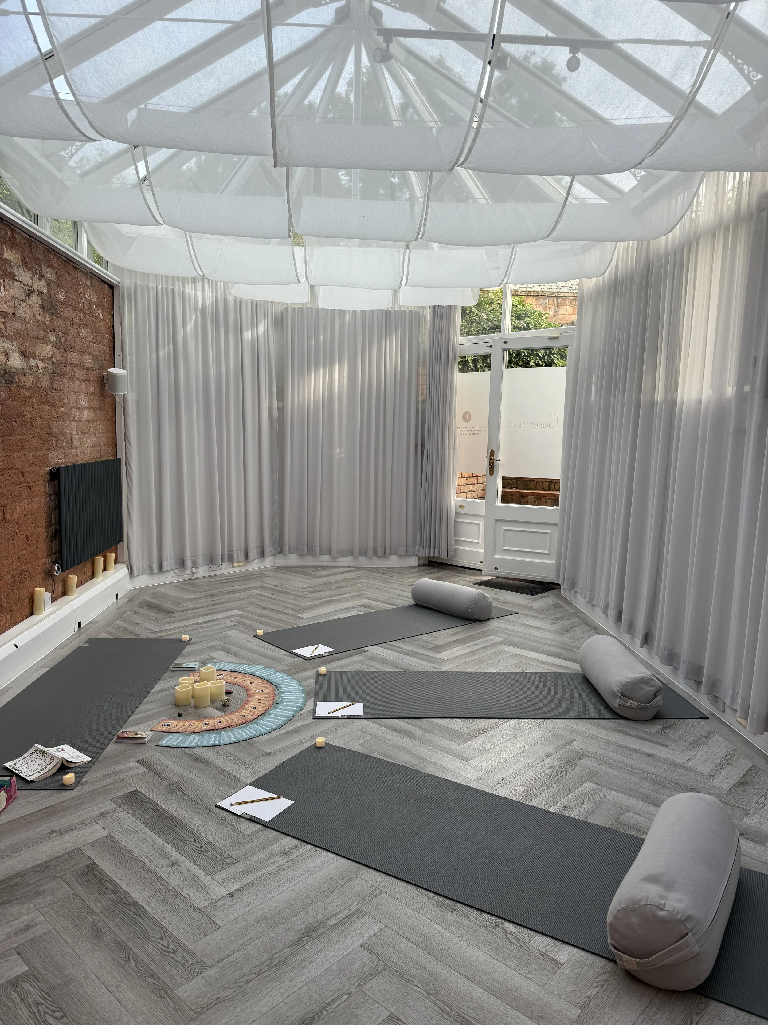 A room set up for yoga or meditation with yoga mats, bolsters, candles, and notebooks on clipboards, with white curtains and a glass ceiling.