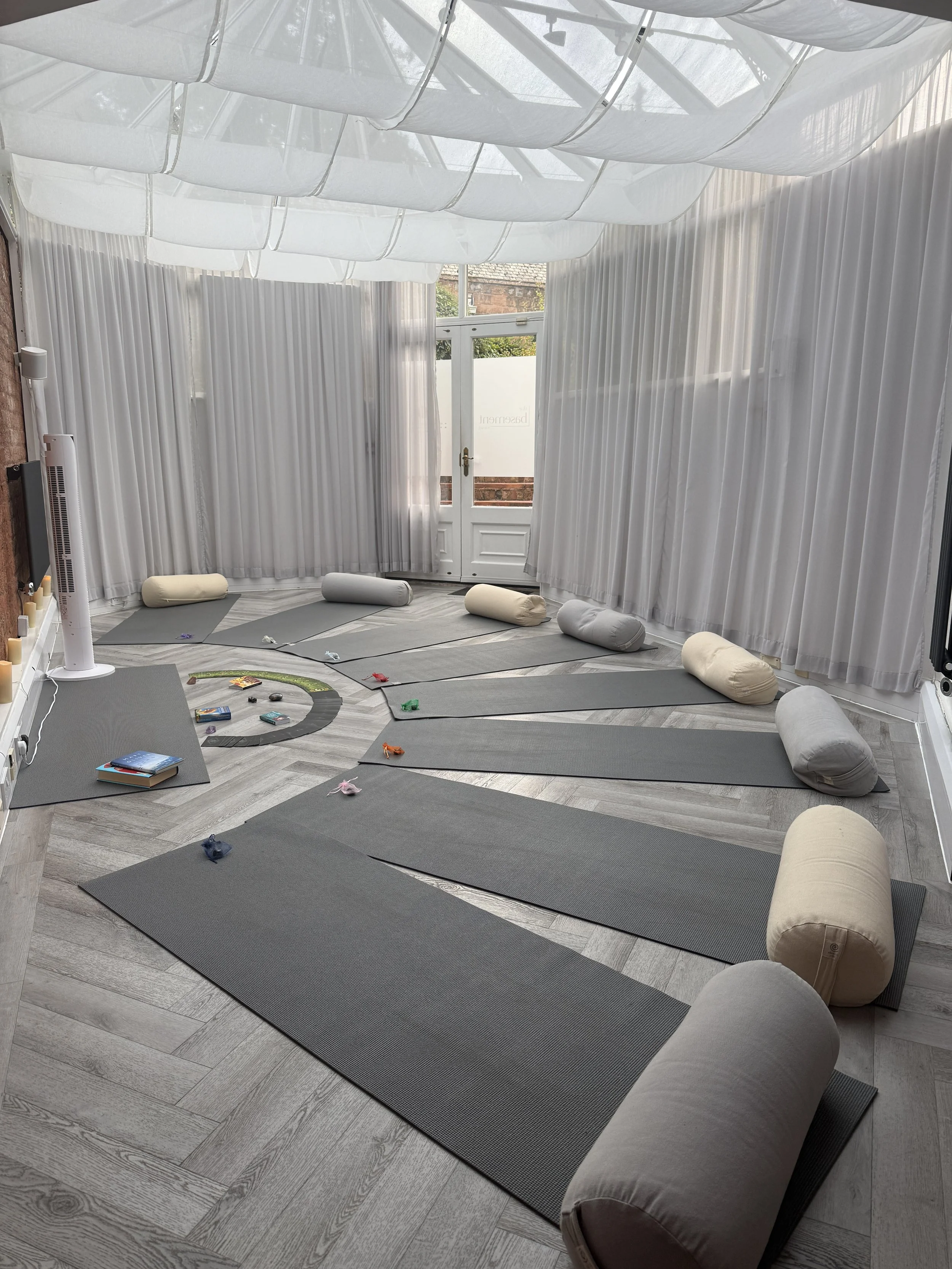 A bright room set up for yoga or meditation with multiple grey yoga mats laid out on a wooden floor, several pillows along the walls, and a glass door leading outside, surrounded by white curtains.