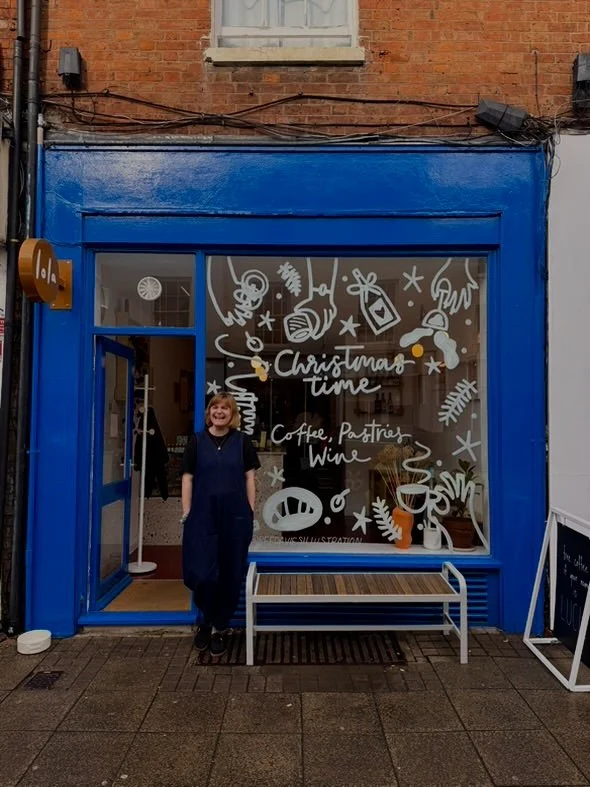 A woman standing in front of a storefront painted bright blue with a large window. The window has Christmas-themed decorations and text reading "Christmas time" and mentions coffee, pastries, and wine. Inside the store, some furniture and plants are visible.