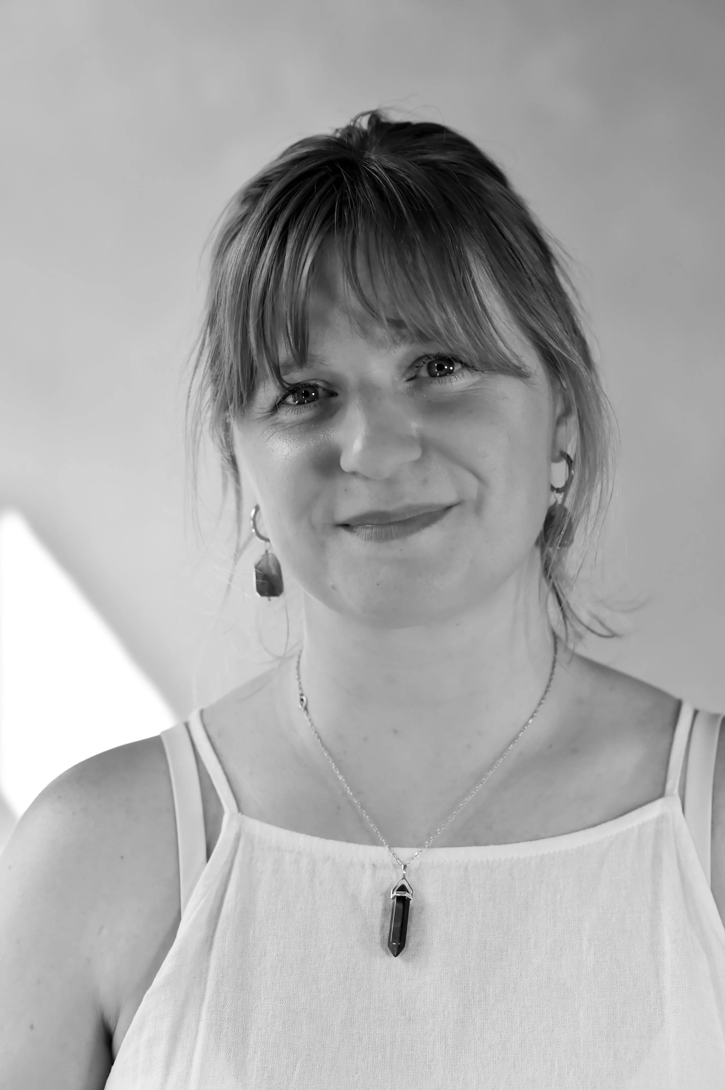 A woman with light-colored hair styled with bangs and earrings, wearing a sleeveless top and a necklace with a large pendant, smiling in a close-up black and white portrait.