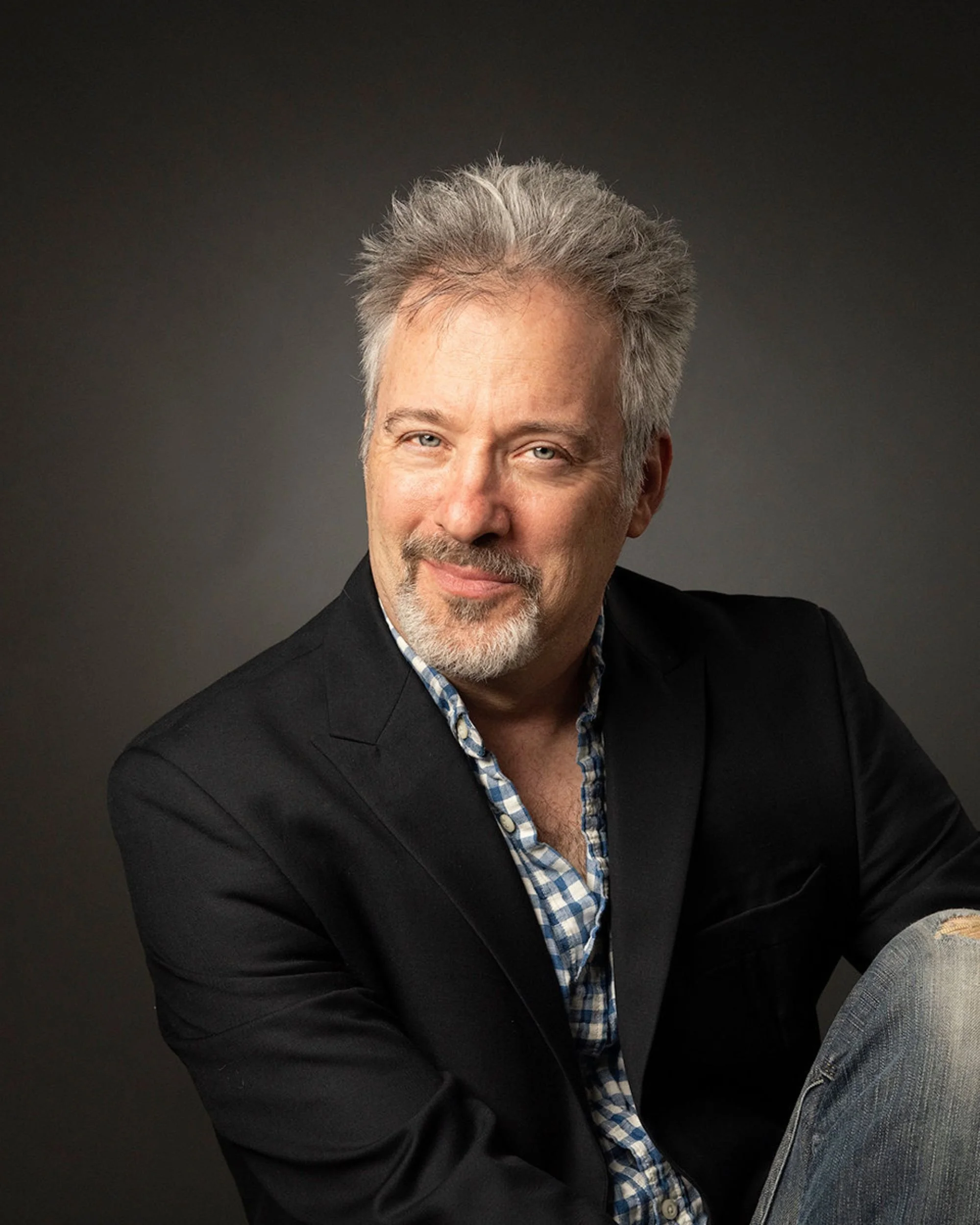 Close-up of an older man with gray hair and blue eyes, wearing a dark blazer and a plaid shirt, smiling against a dark gray background.