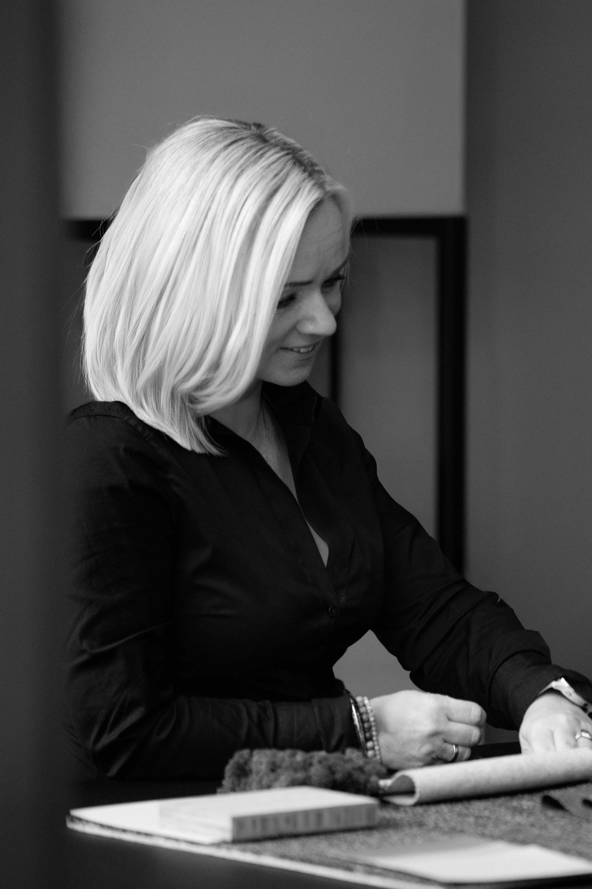 En kvinne med blondt hår som smiler, iført svart skjorte, sitter ved et bord med papir og bøker.