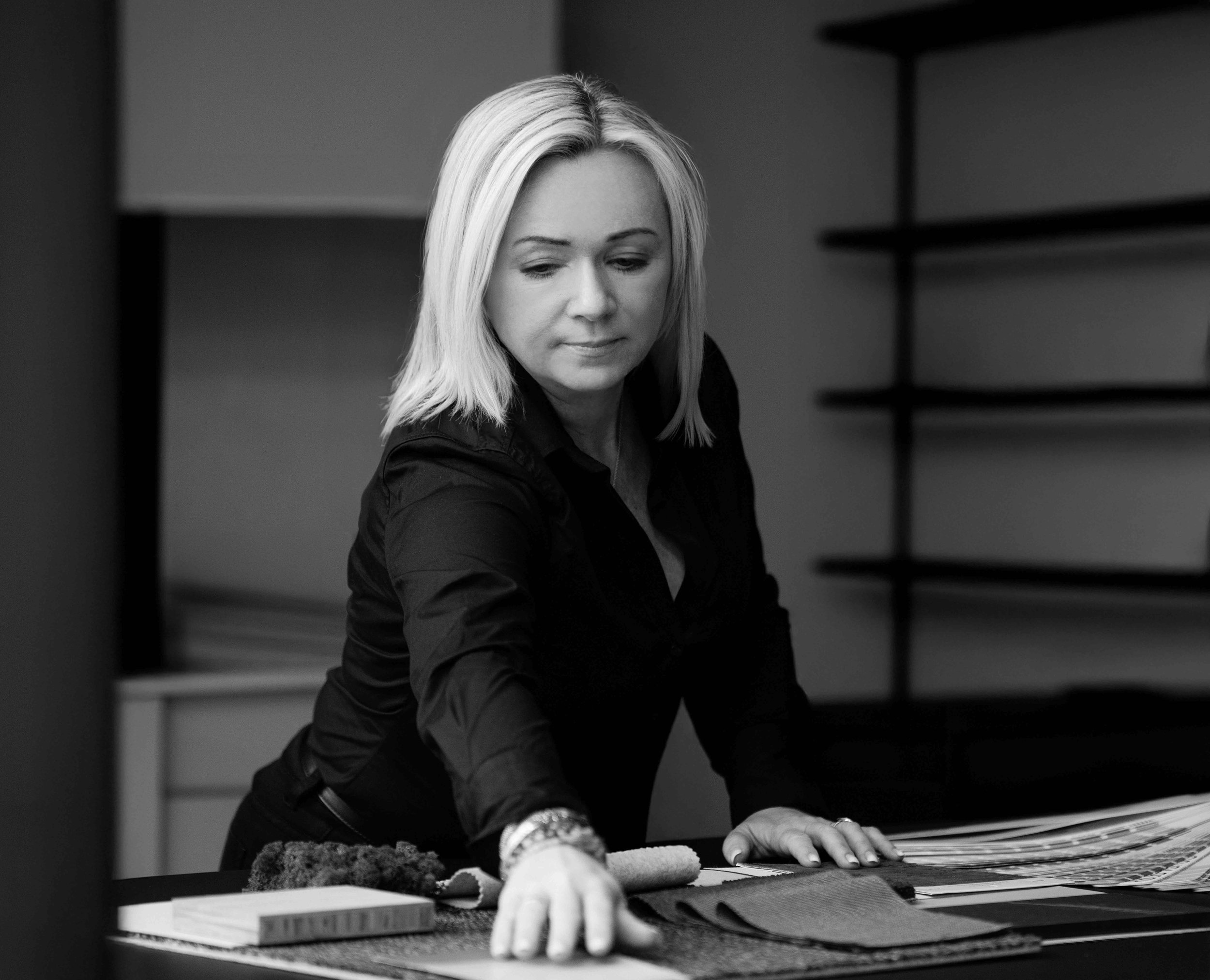 En kvinne med blondt hår i mørk skjorte som arbeider med stoffprøver og fargekart på et bord.