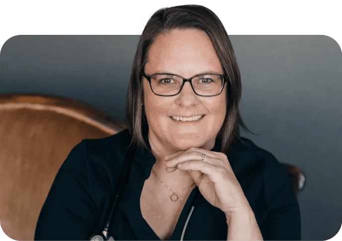 A woman with glasses, short brown hair, wearing a dark blue shirt, and a stethoscope around her neck, smiling and resting her chin on her hand.