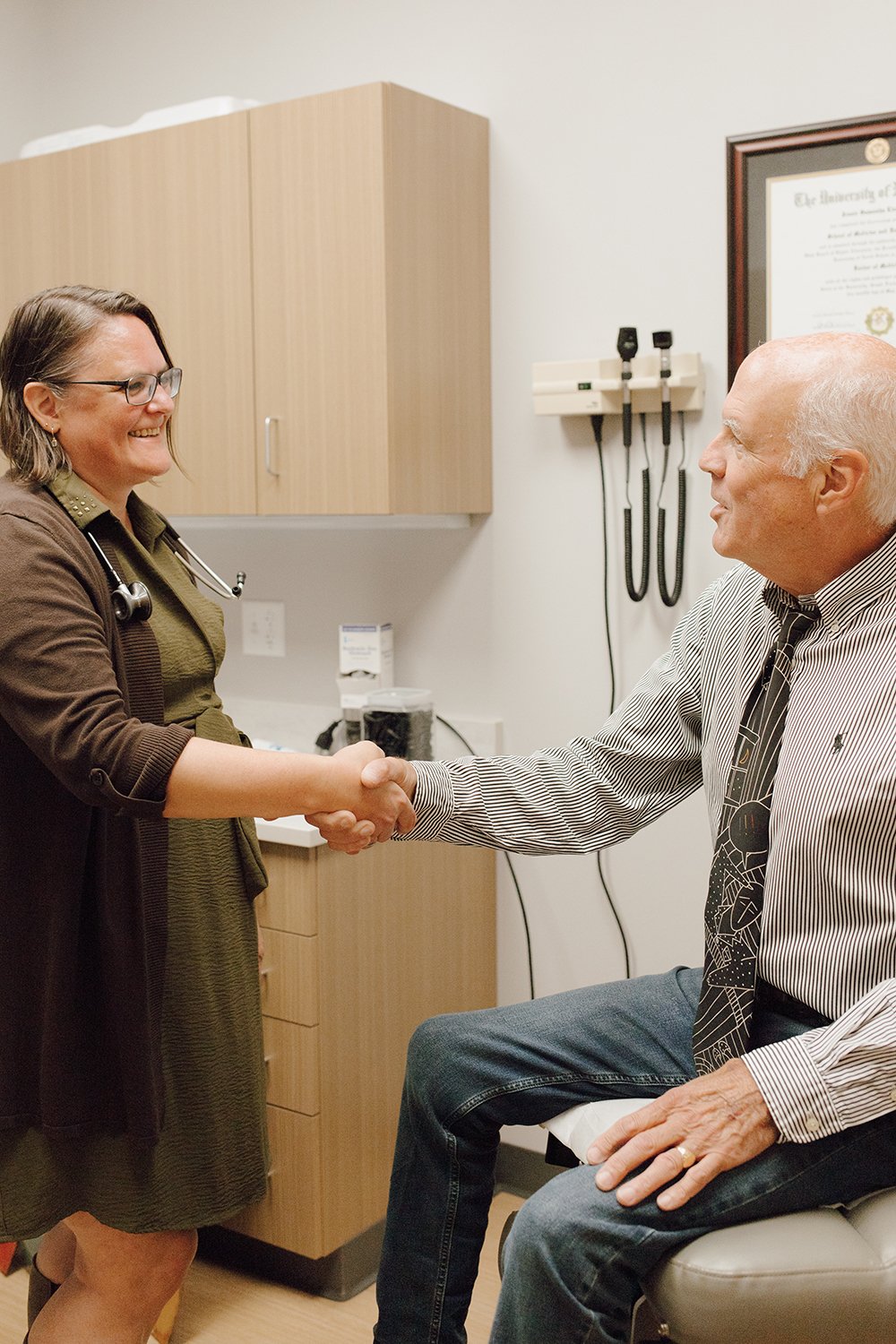 A female doctor or nurse shaking hands with a male patient who is sitting on an examination table, in a medical office or clinic.