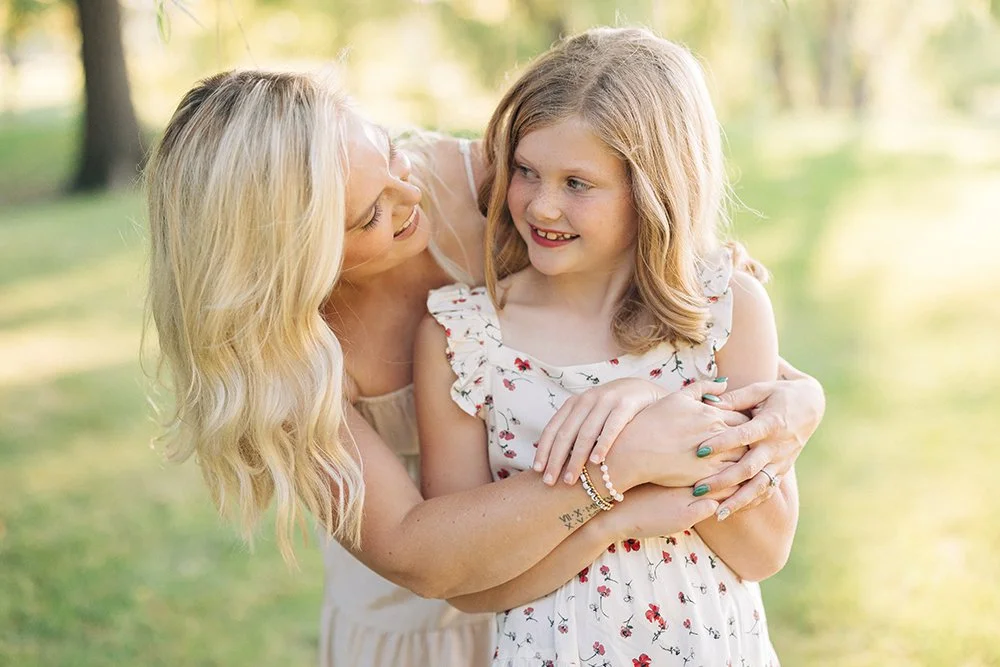 A woman hugging a young girl in a park, both smiling and looking at each other.