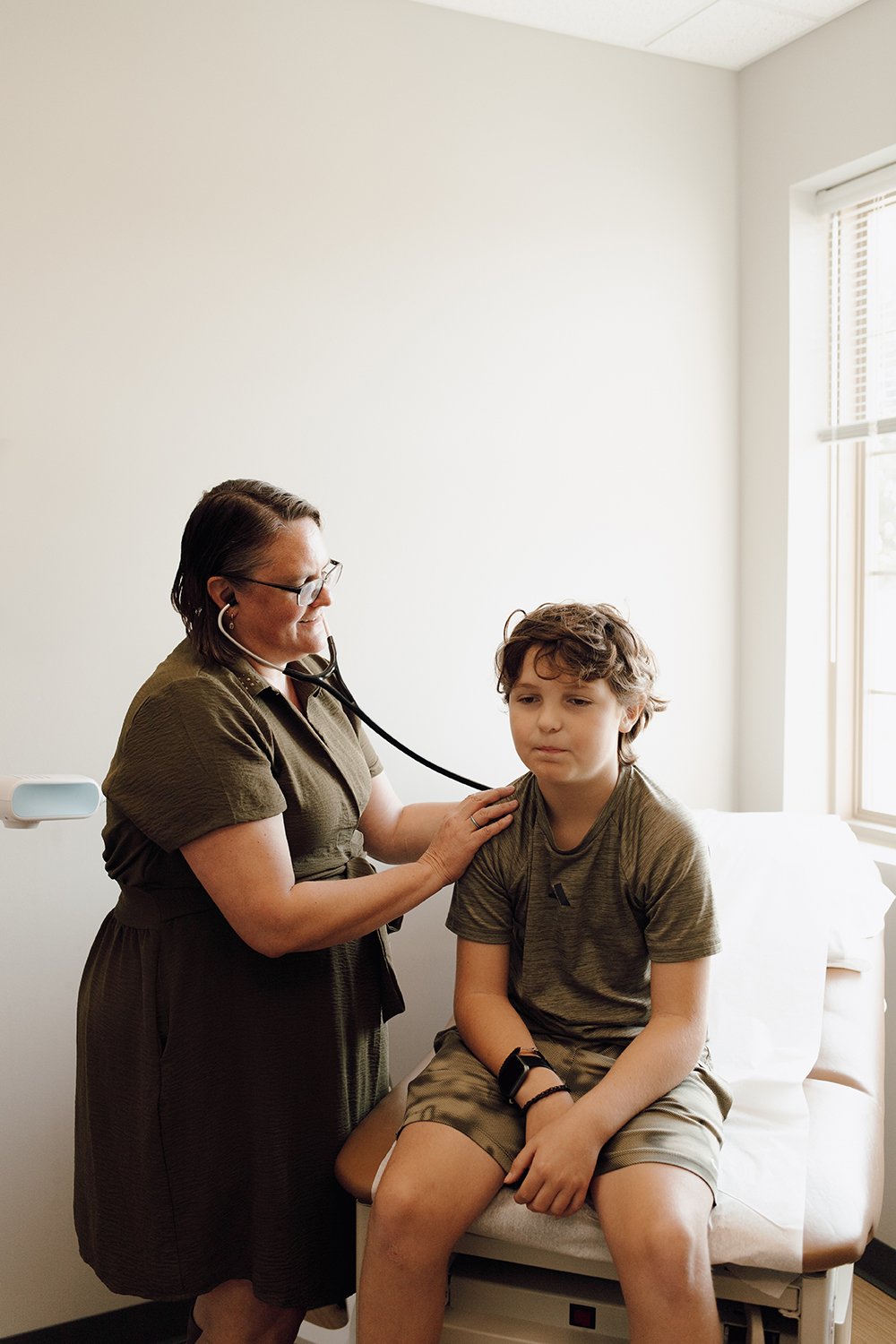 A female healthcare provider using a stethoscope to listen to a young boy's chest during a check-up in a clinic room with a large window.