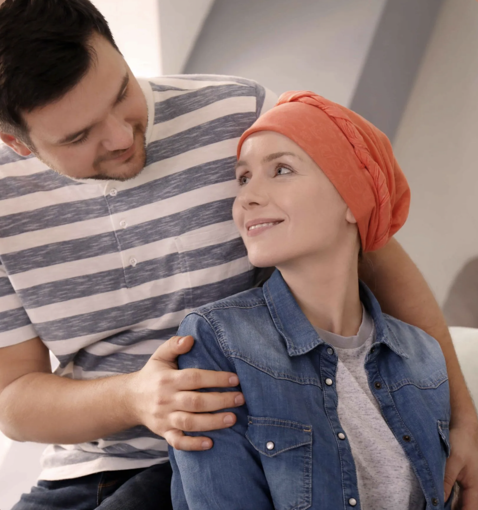 A man and woman share a tender moment indoors, the woman wearing a red headscarf and denim jacket, smiling at the man, who has his hand on her shoulder and looks at her affectionately.