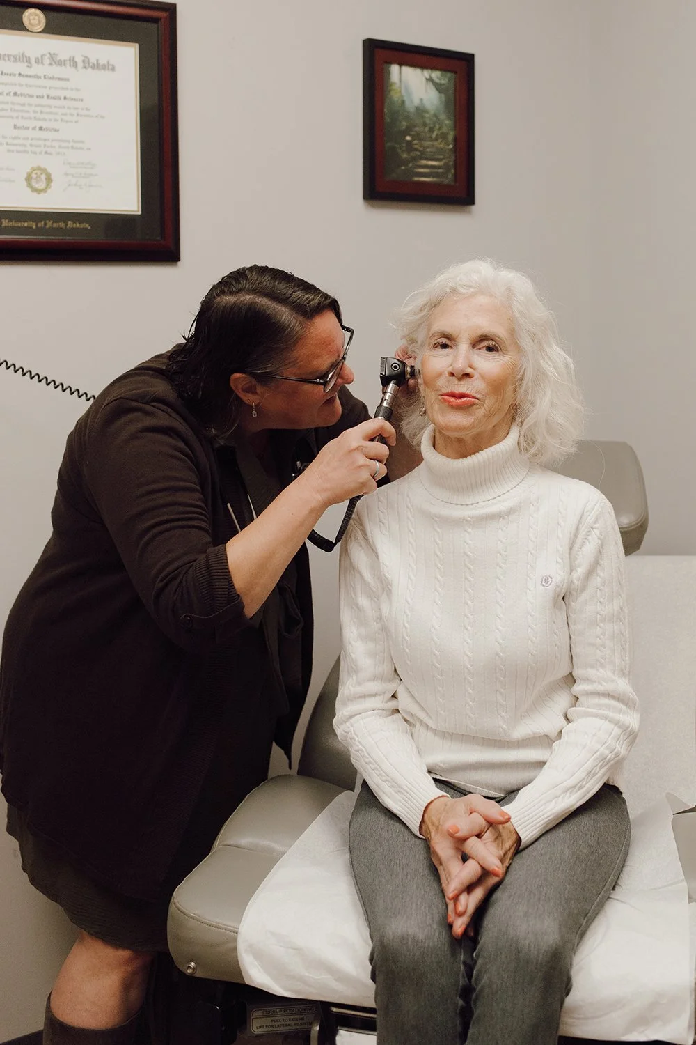 A woman with white hair sitting on an examination table while a healthcare professional examines her ear with an otoscope. The woman is wearing a white turtleneck sweater and gray pants. The healthcare professional is leaning in, wearing glasses and a dark jacket. There are framed certificates and pictures hanging on the wall behind them.