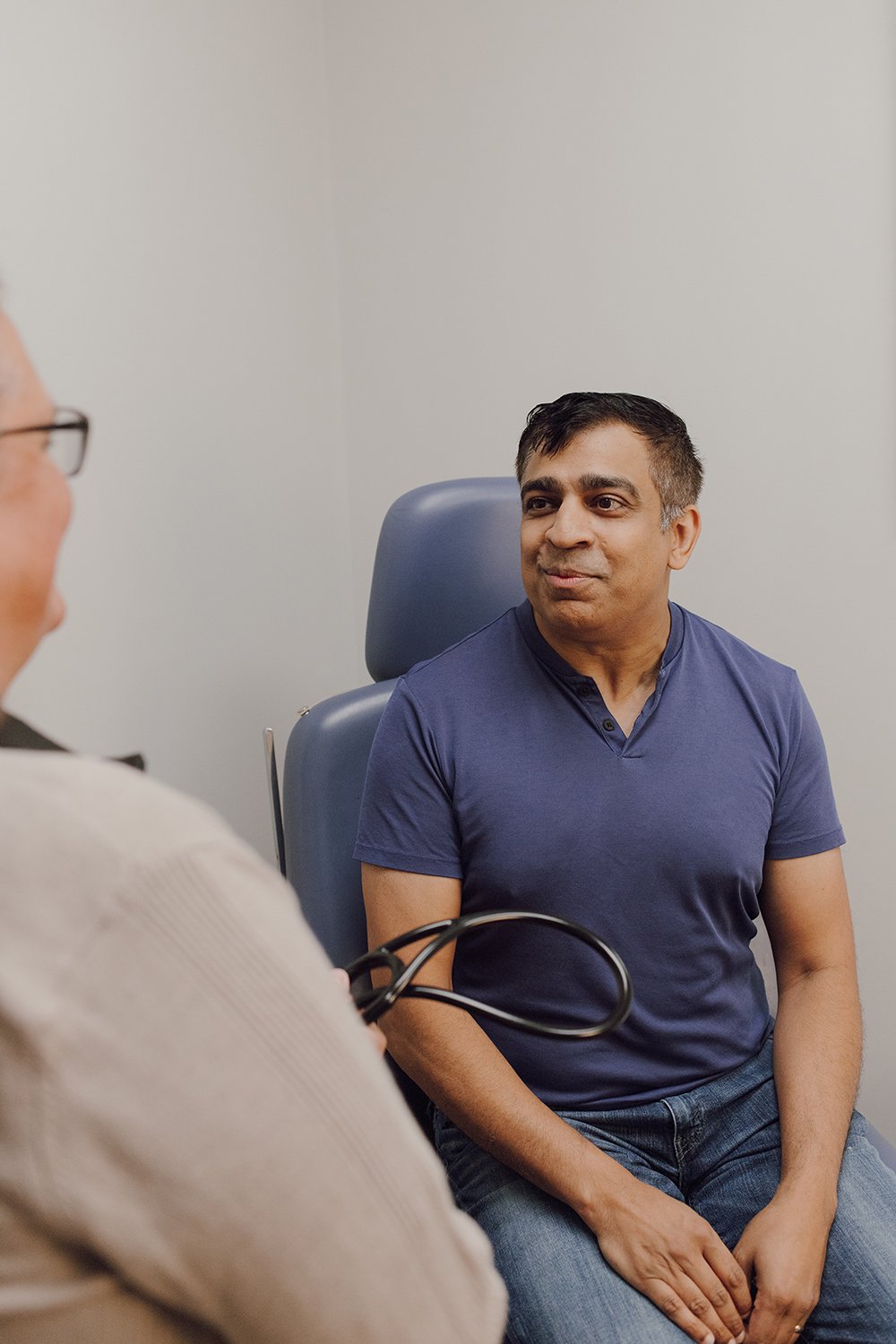 A man sitting in a doctor's office, talking to a doctor who is partially visible, with a stethoscope in the foreground.