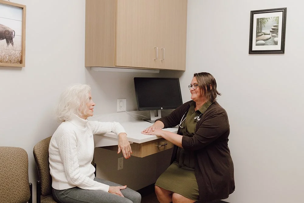 A healthcare professional talking to an elderly woman in a medical office, with a computer on the desk and framed pictures on the walls.