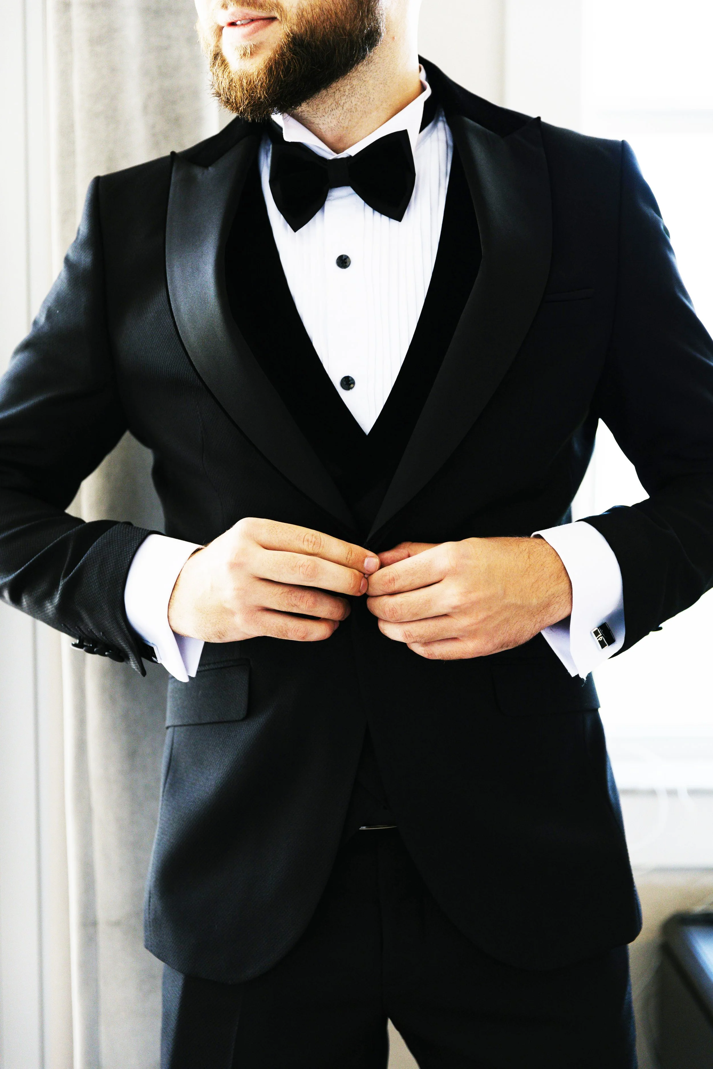 A man in a black tuxedo with a bow tie is buttoning his jacket, standing indoors near a window.