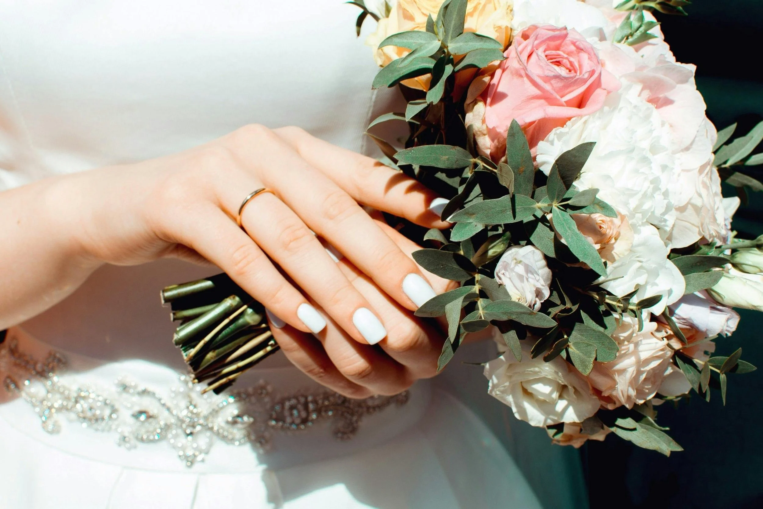 Close-up of a woman's hand with a wedding ring holding a bouquet of pink and white roses, green leaves, and eucalyptus, with part of a white dress and jewelry visible.