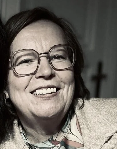 Frau mit Brille, lächelnd, in einem Raum mit unscharfem Hintergrund.