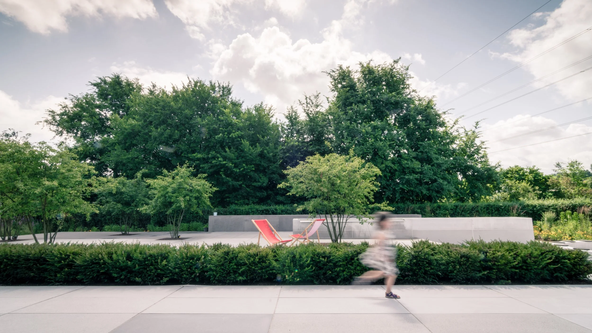 A young boy running on a sidewalk with green bushes and trees in the background on a sunny day with some clouds.