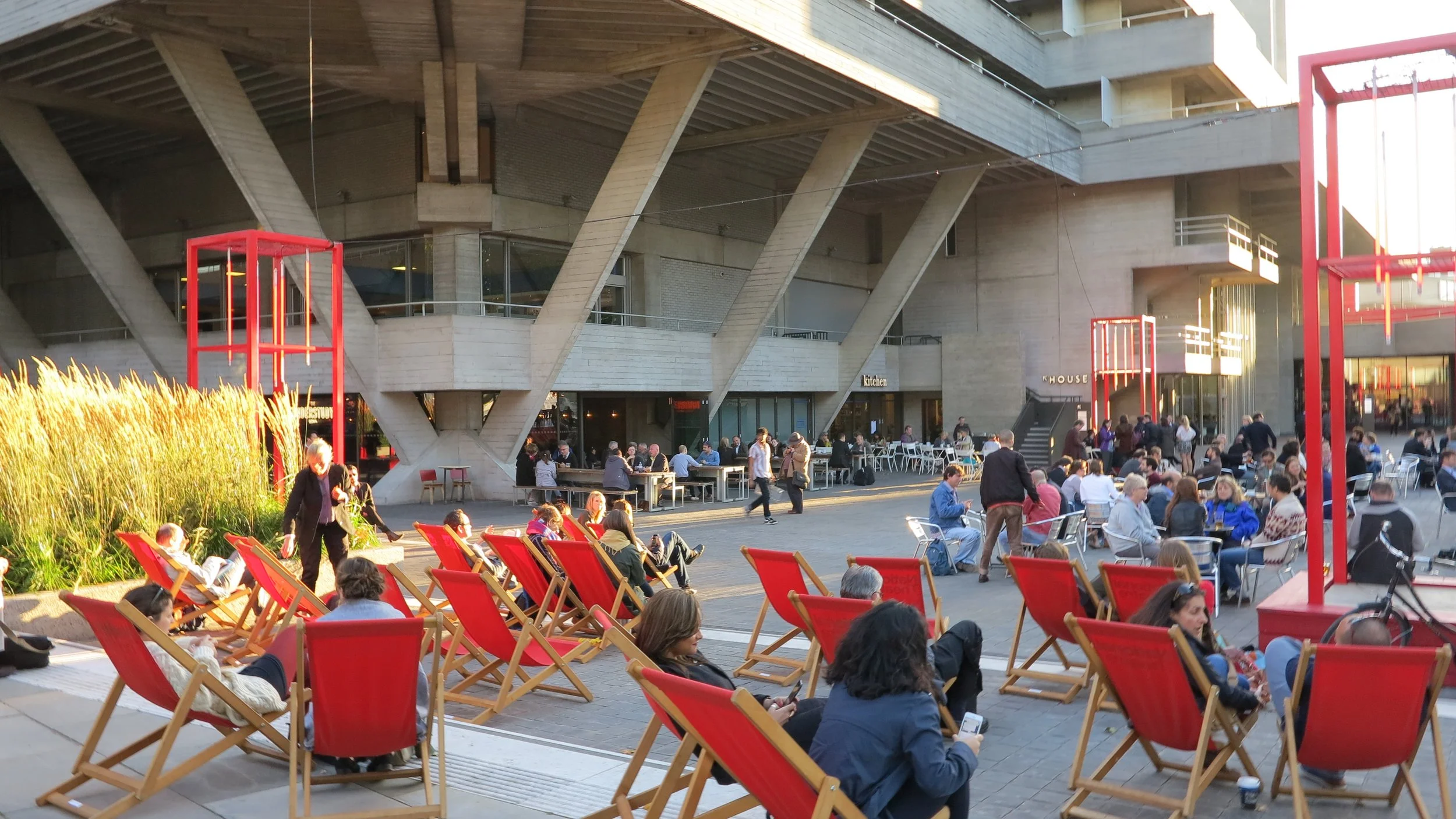 People relaxing on red deck chairs in an outdoor urban plaza with a modern concrete building in the background