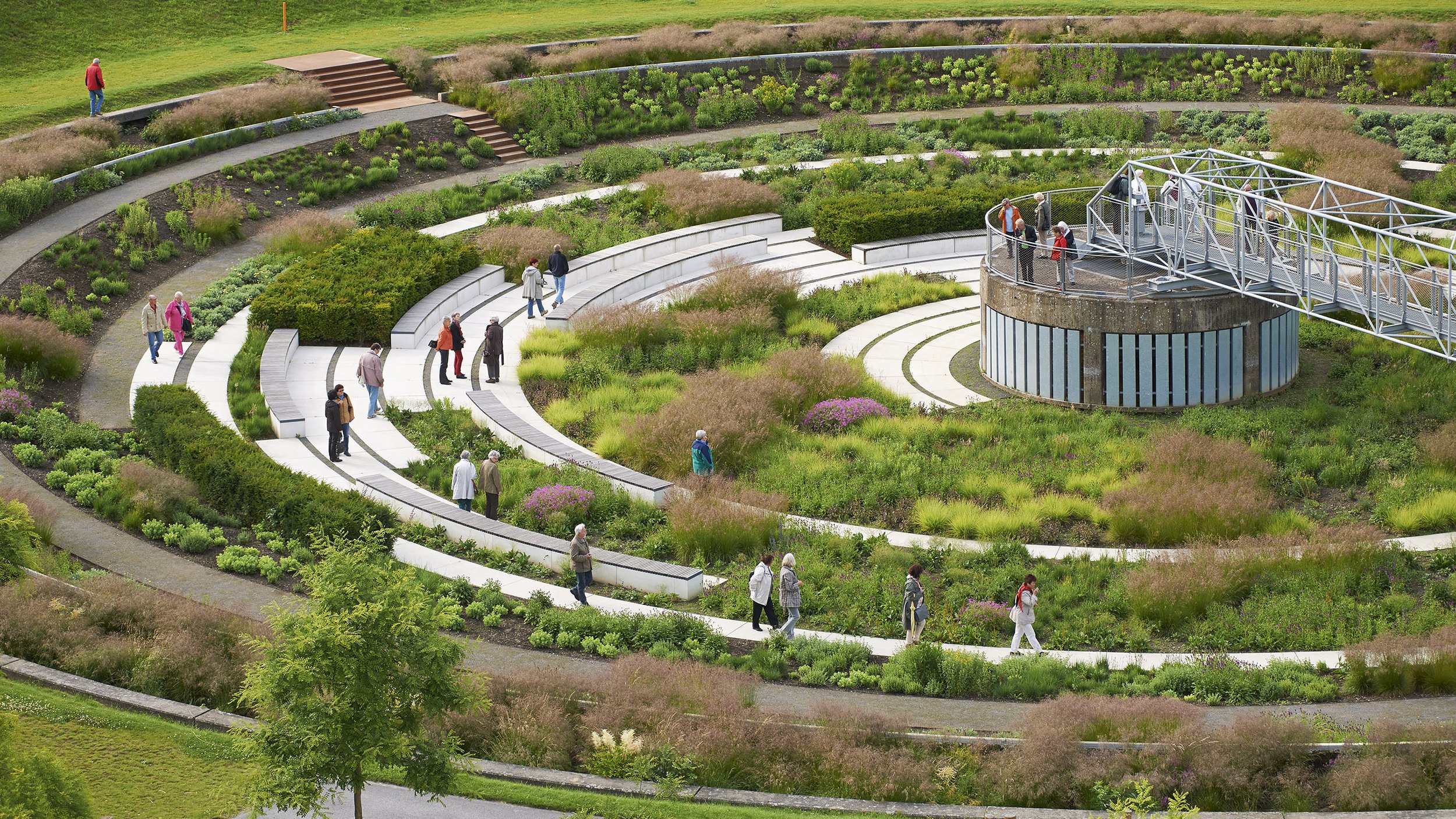 A spiral-shaped outdoor amphitheater with concrete steps, surrounded by landscaped greenery and people walking along the paths. There is a round, elevated platform with a metal walkway and a group of people on it.