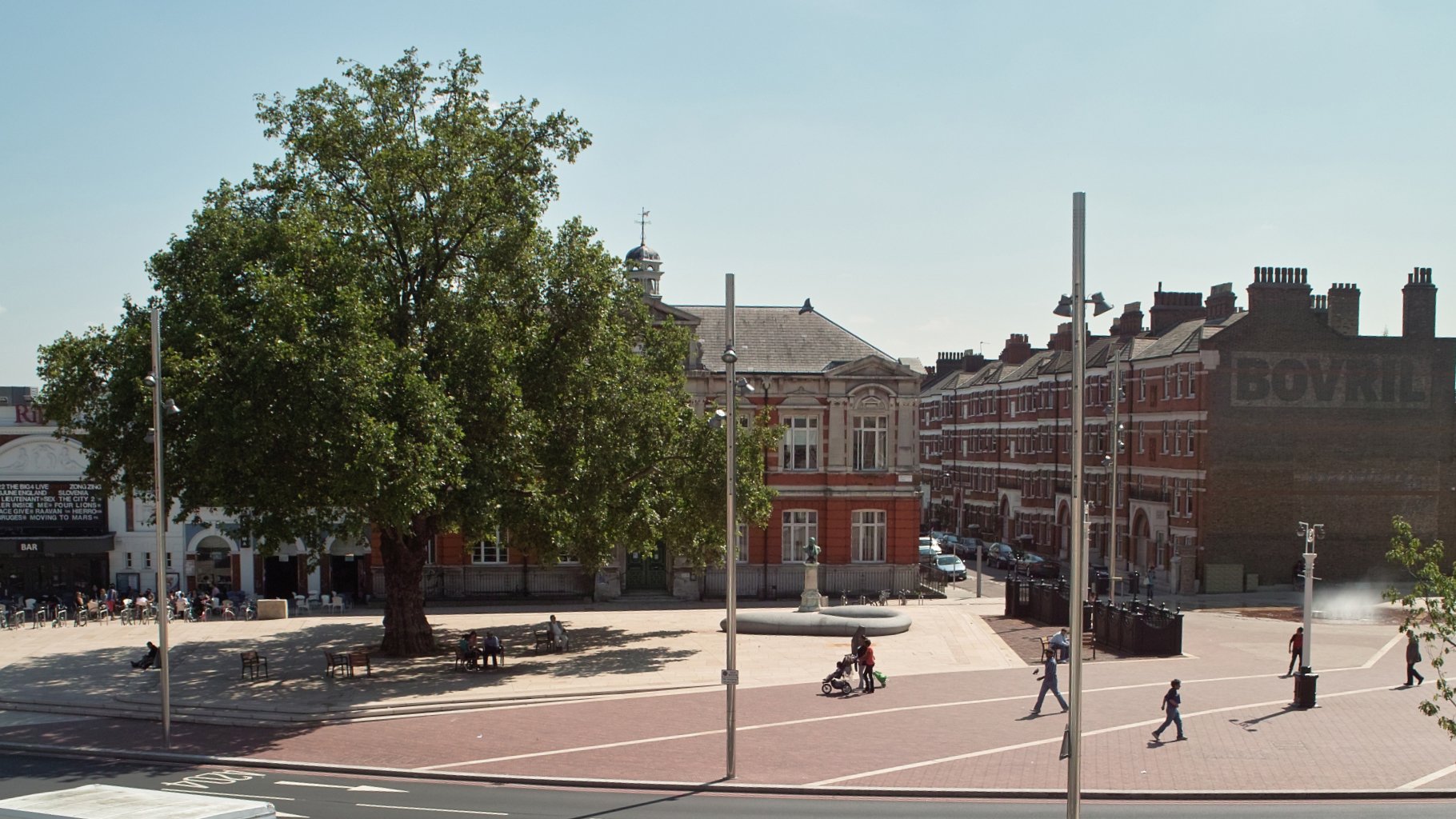 A park with a large tree, benches, people walking and sitting, and historic buildings in the background on a sunny day.