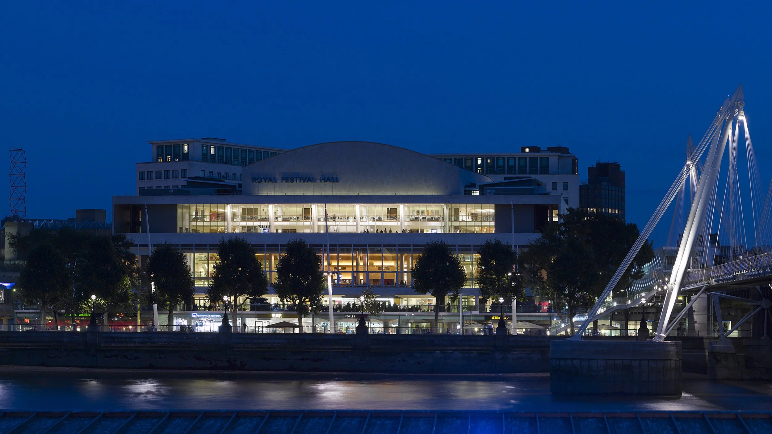 Night view of the Royal Festival Hall with illuminated windows, trees in front, and a modern bridge to the right, reflecting on the river.