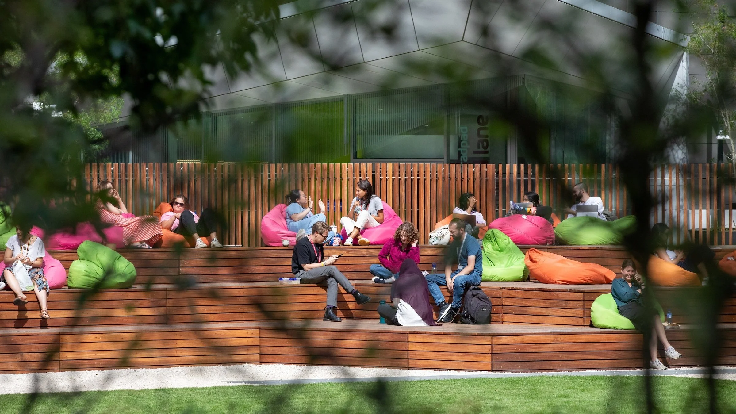People sitting on wooden steps with colorful bean bags, some chatting, some using laptops or phones, in an outdoor urban space surrounded by greenery and a tall wooden fence.